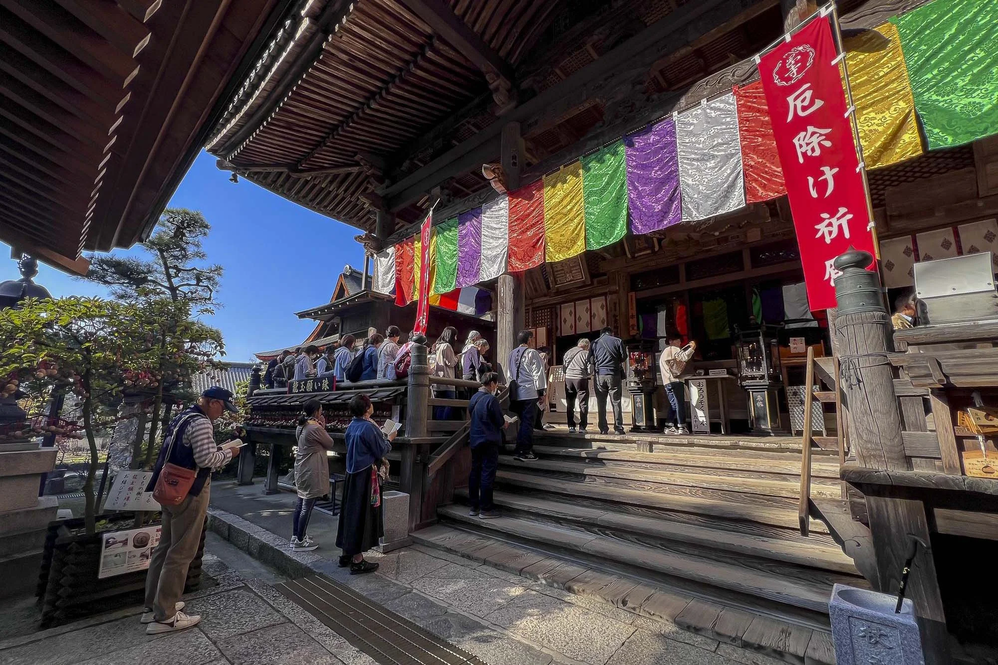 People waiting in line at the entrance of a traditional Japanese temple, with colorful banners hanging overhead and a clear blue sky in the background.
