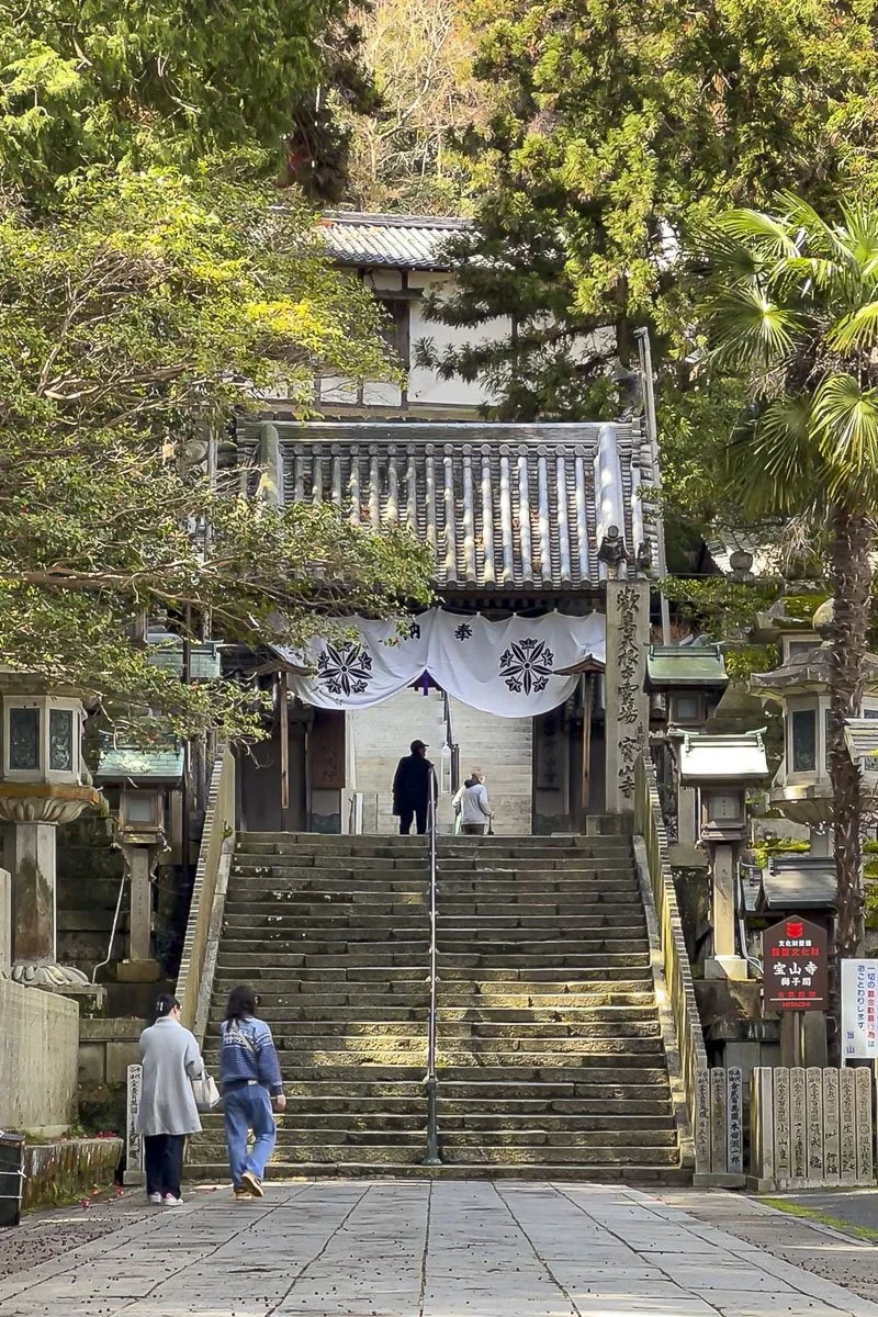 Stone steps leading up to a traditional Japanese shrine entrance, surrounded by trees and lanterns.