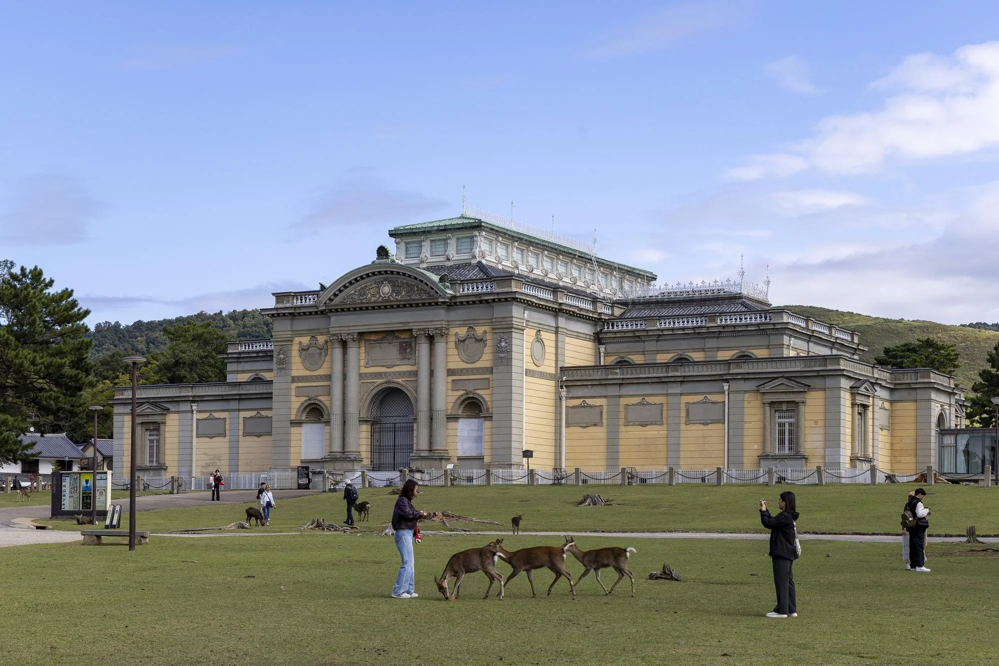 People walking and taking photos of deer in front of a historic, ornate building on a grassy lawn with trees and hills in the background.