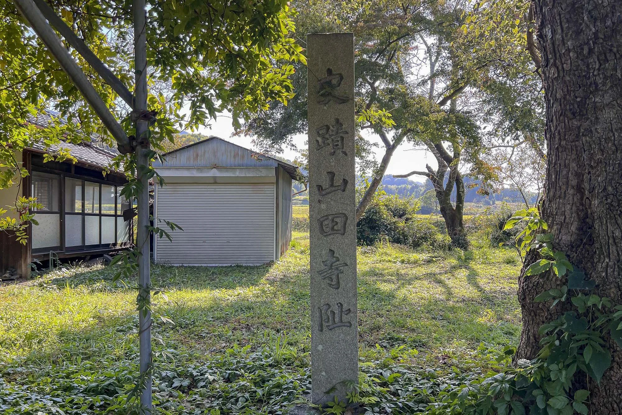 A stone monument with Japanese characters stands among trees and greenery in a rural area, with a small wooden building and trees in the background.