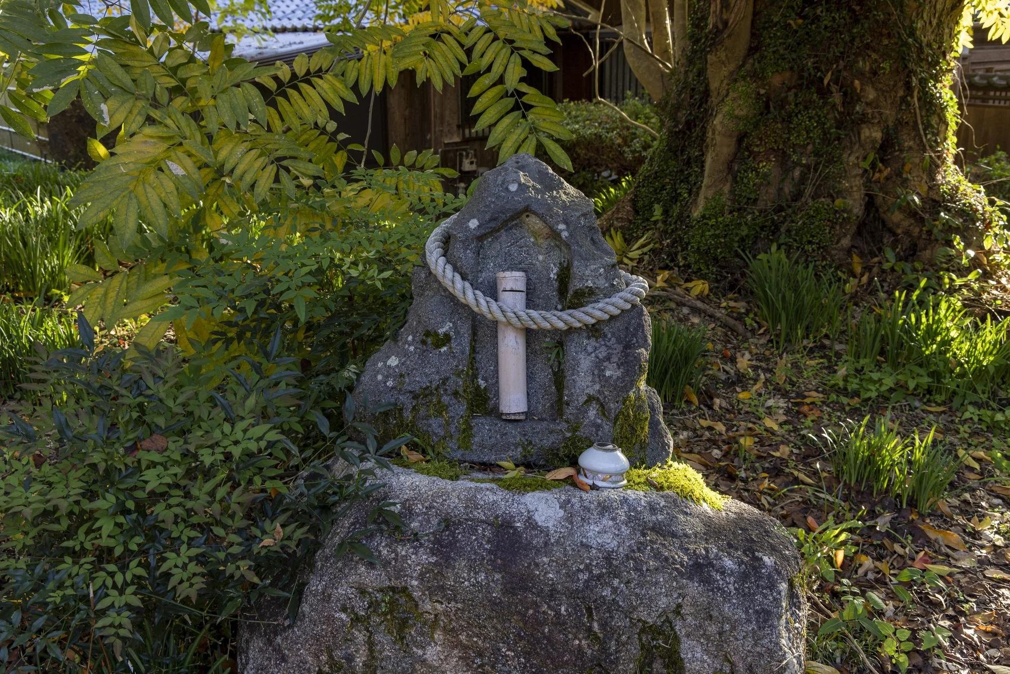 A small stone shrine with a white rope tie, a wooden cross, and a small ceramic container is situated on a large moss-covered rock in a garden, surrounded by green foliage and trees.