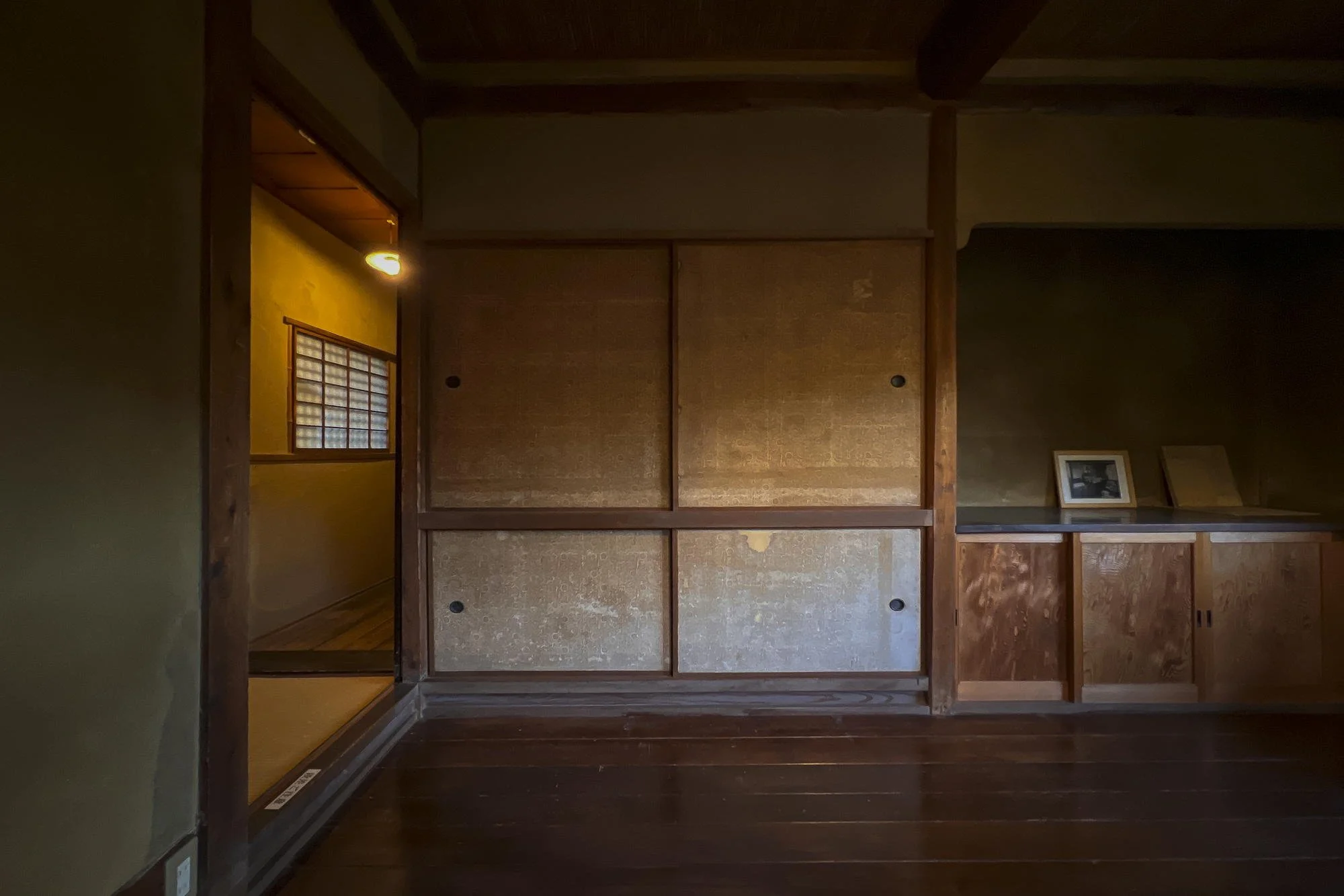 Empty traditional Japanese room with sliding wooden doors, tatami mats, and framed photographs on a dark wooden shelf.