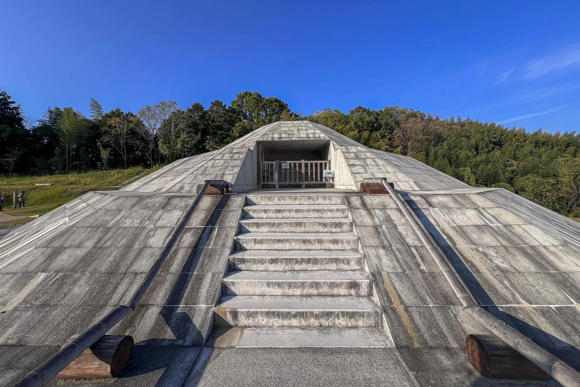 View of the roof of the Tsunami Museum in Japan, with stairs and rails leading up to an entrance, surrounded by trees and a clear blue sky.