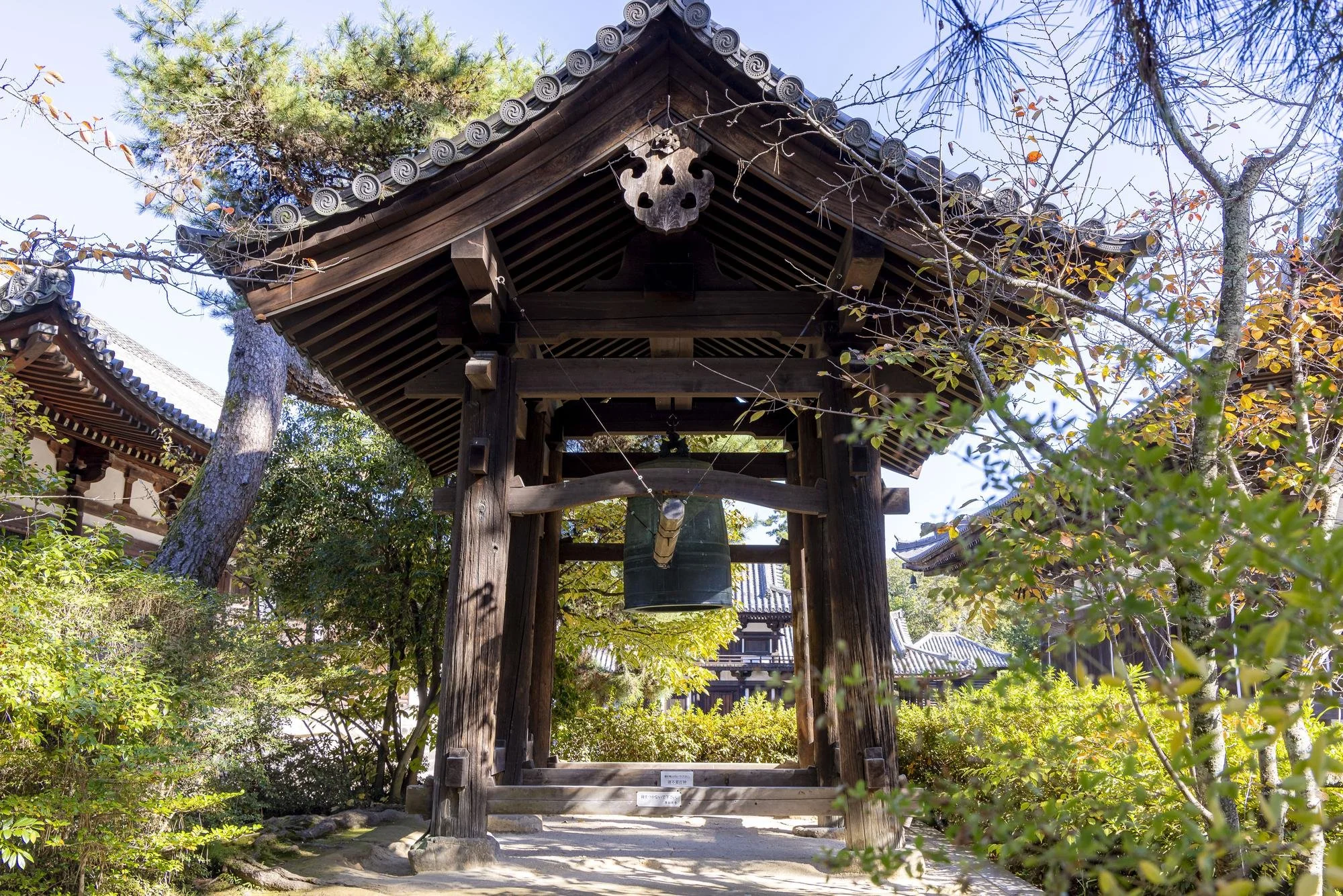 Traditional Japanese temple bell tower with a large hanging bell, surrounded by trees and shrubs, with a clear sky in the background.