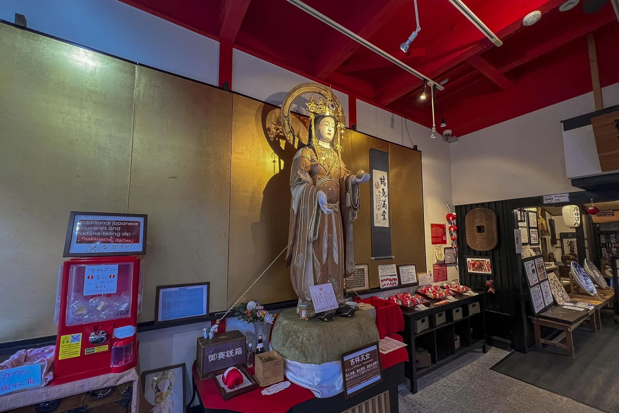 A Buddhist or Shinto religious statue of a female deity or bodhisattva, with intricate robes, a halo, and jewelry, standing on a pedestal inside a shrine with various displays, signs, and red decorations around.