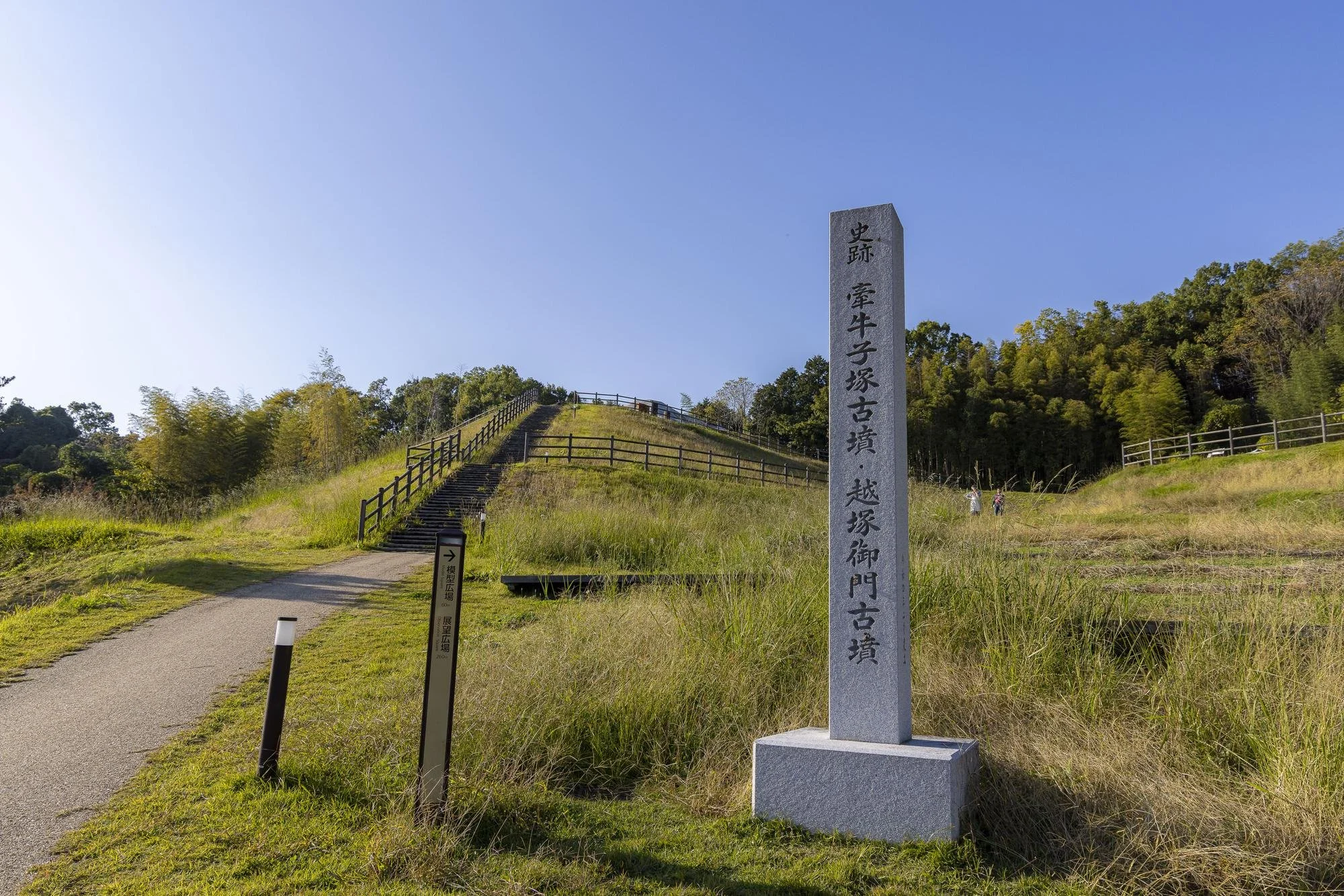 A stone monument with Japanese inscriptions on a grassy area near a path and a staircase, with trees and a clear blue sky in the background.