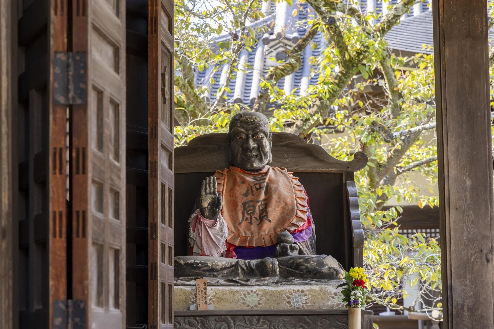 Statue of a seated Buddhist monk or deity inside a temple, with a raised right hand in a blessing gesture, dressed in a pink and purple robe, with Japanese characters on a cloth around its neck, and a flower arrangement in front, surrounded by wooded