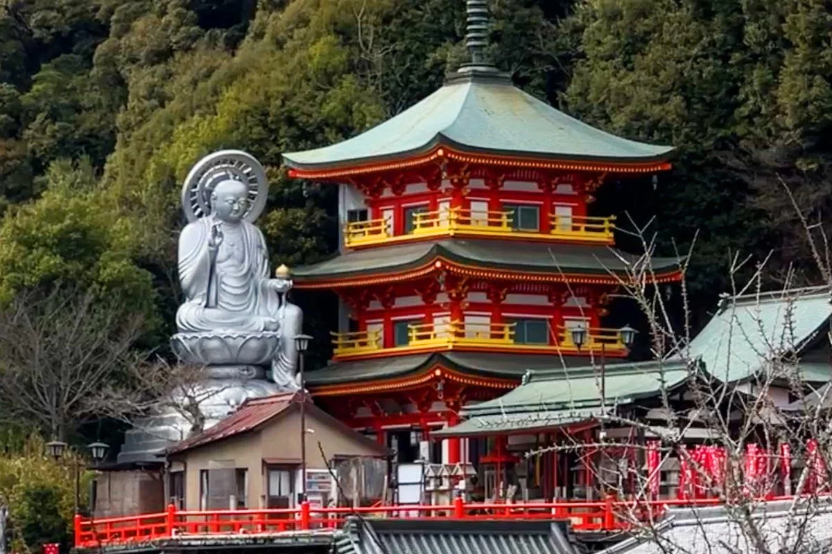 Japanese temple with a large silver Buddha statue, red and yellow accents, and a green roof, surrounded by trees and located near a hillside.