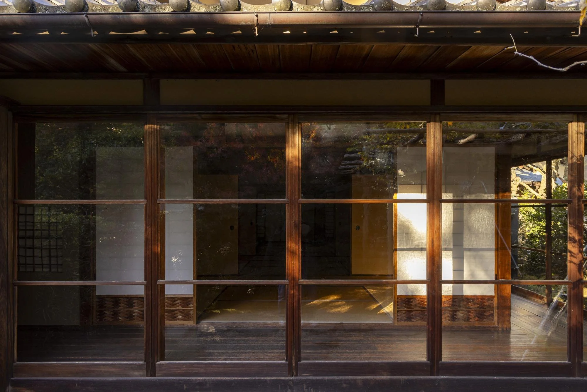 Wood-framed glass wall of a house or room, with visible interior wooden floors and some furniture, and reflections of trees and the outdoors on the glass.