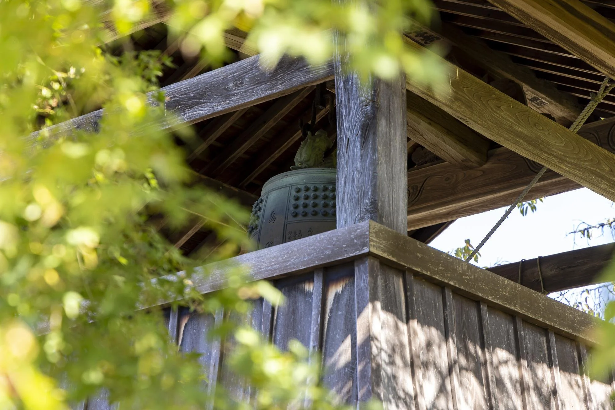 A wooden bell tower with a large, decorative bell inside, partially obscured by green foliage, showing the rustic wood structure and roof.