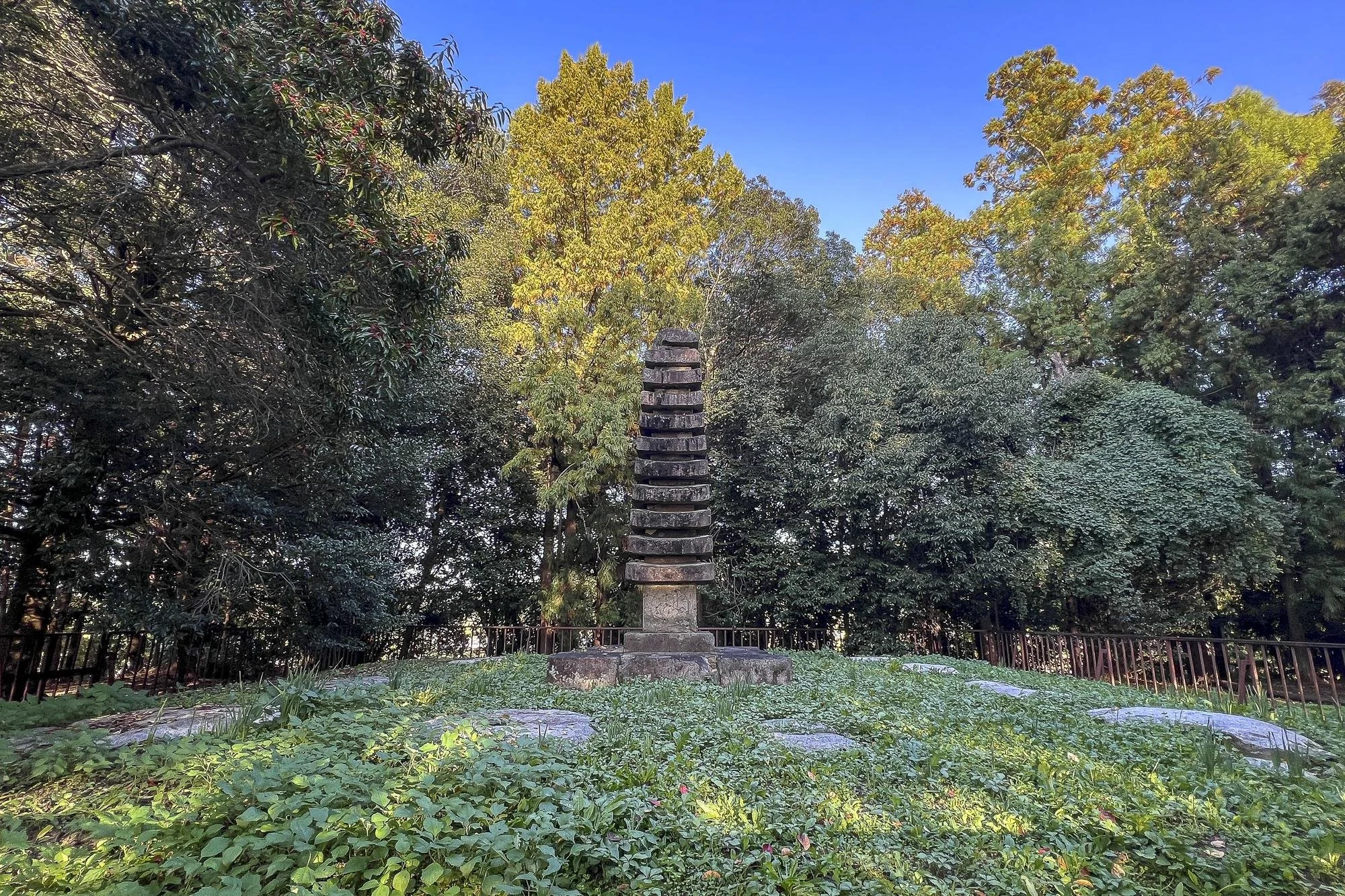 Hinokuma-dera Temple Site: A wooden pagoda-style sculpture on a stone base in a grassy area surrounded by dense green trees like oaks and pines under a clear blue sky.