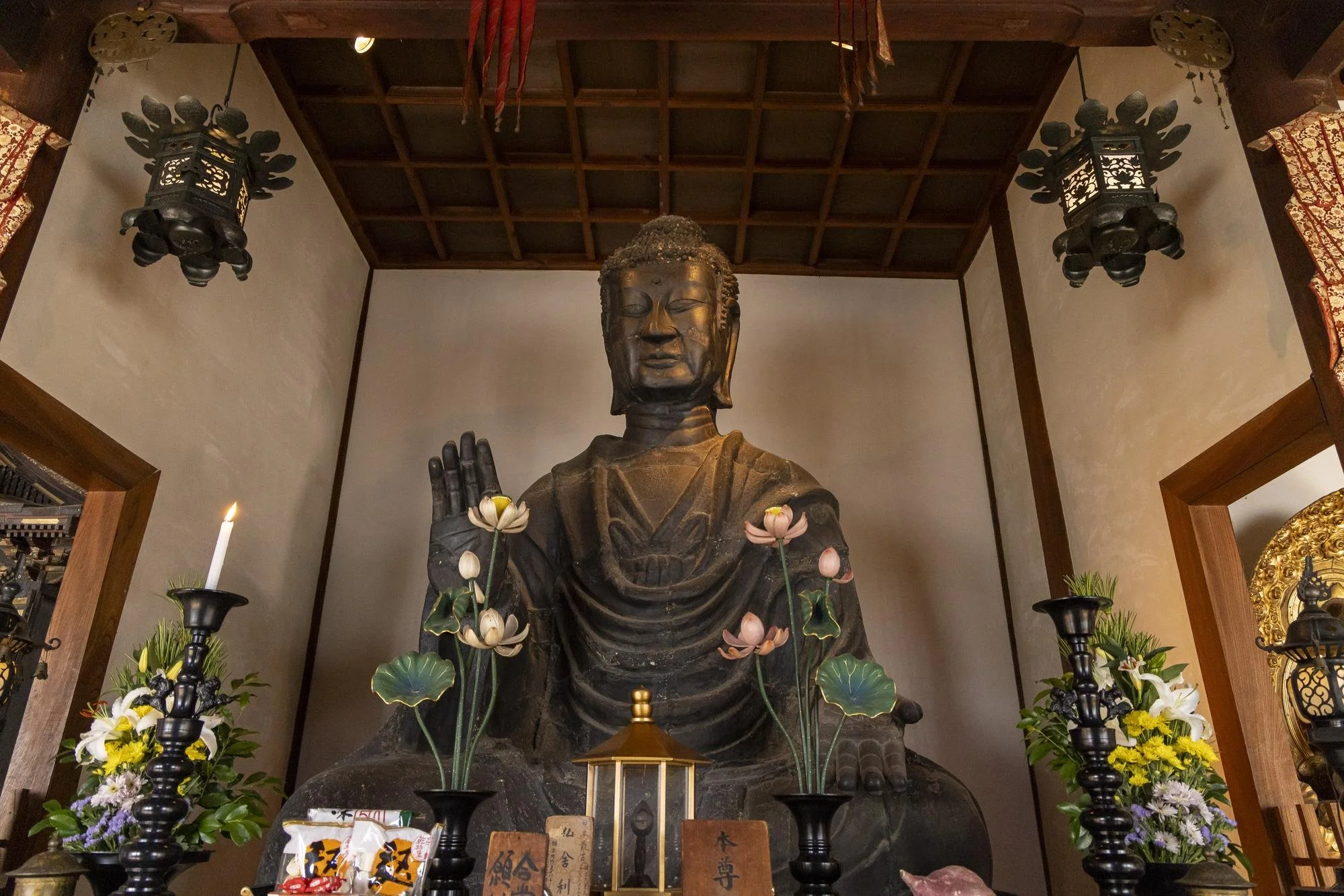 A large seated Buddha statue inside a temple, surrounded by flowers, candles, and offerings, with a wooden ceiling and lanterns overhead.