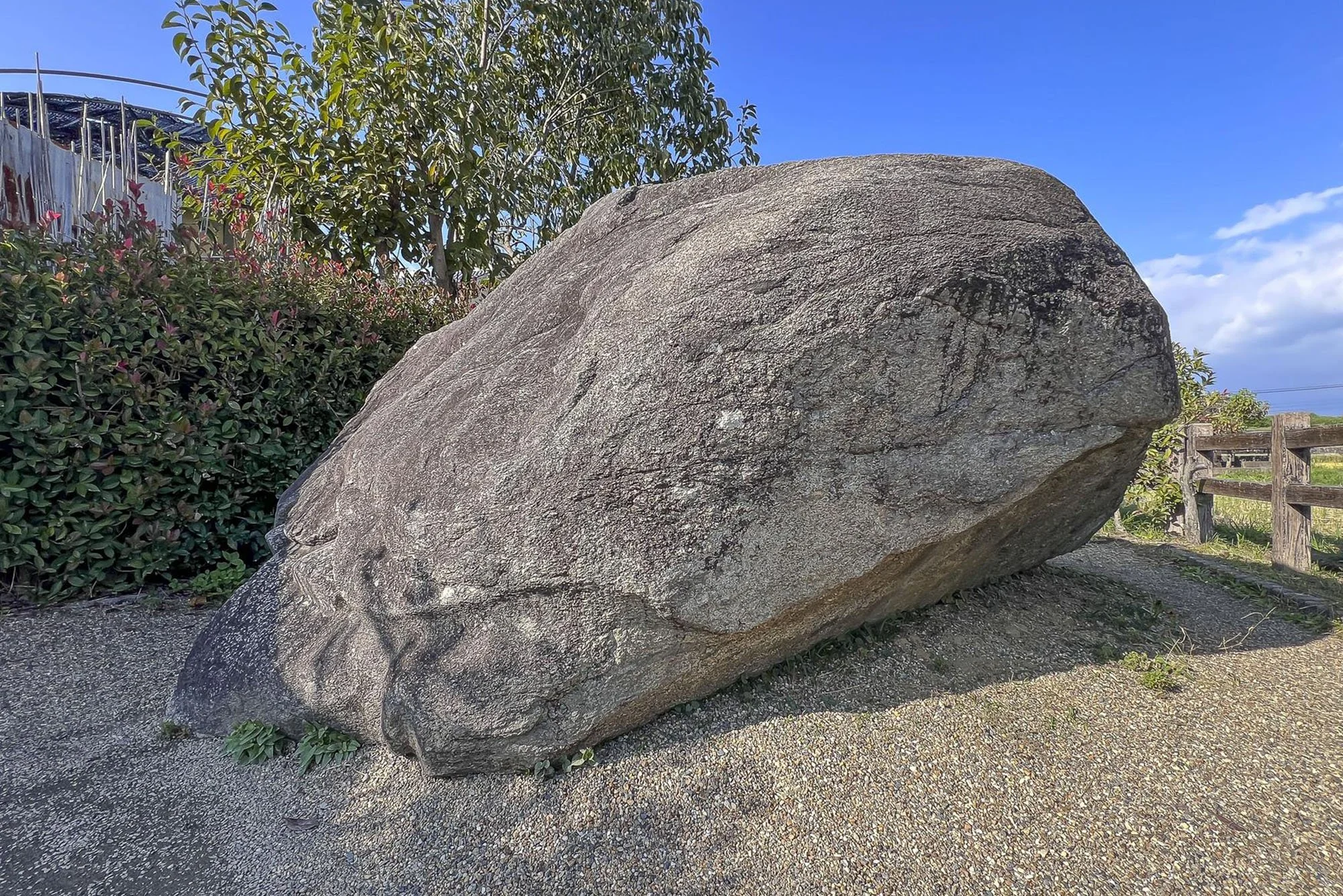 Large gray boulder on gravel path with bushes, trees, and a wooden fence in the background under a blue sky with clouds.