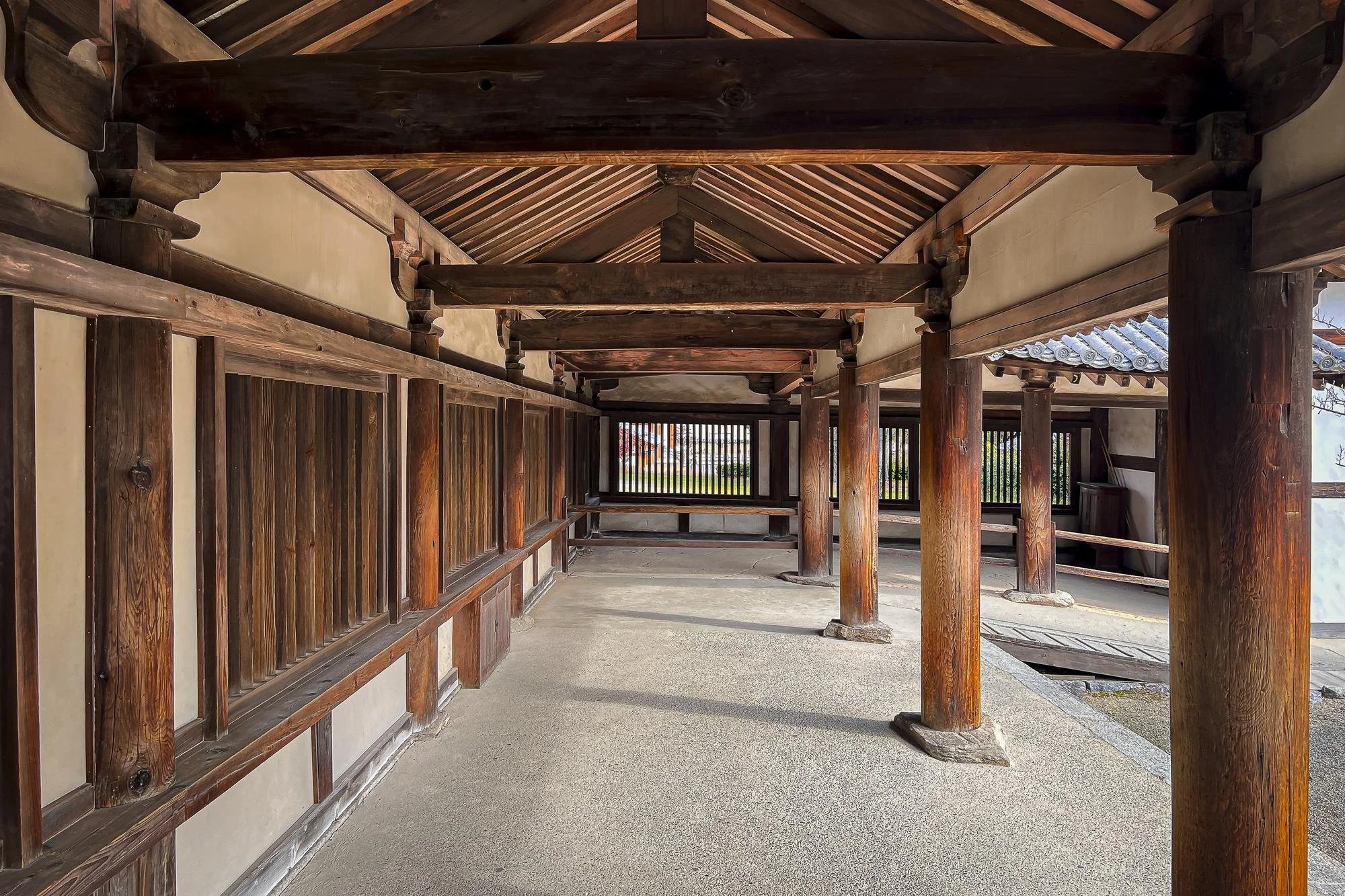 Traditional wooden structure with wooden beams, pillars, and latticework, situated on a smooth concrete floor with partial view of a tiled roof on the right and open space beyond.
