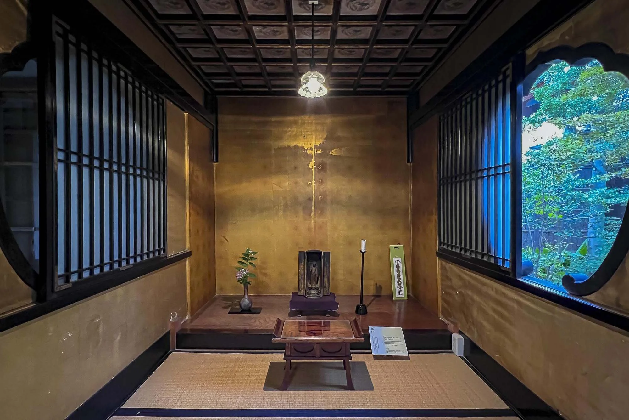 A small traditional Japanese room with a central altar featuring a religious artifact, flanked by a flower arrangement and candle holder, with sliding shoji-style windows on either side revealing a garden view.