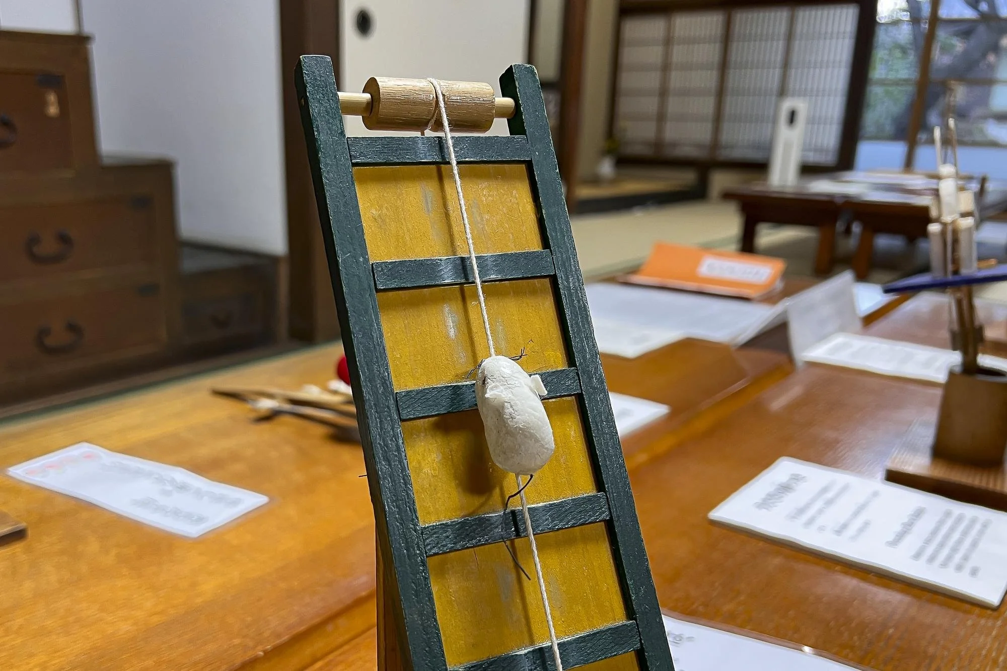 A model of a small white rodent made of papier-mâché hanging on a string attached to a wooden structure with black and yellow colors, in a room with tables, papers, and wooden furniture.