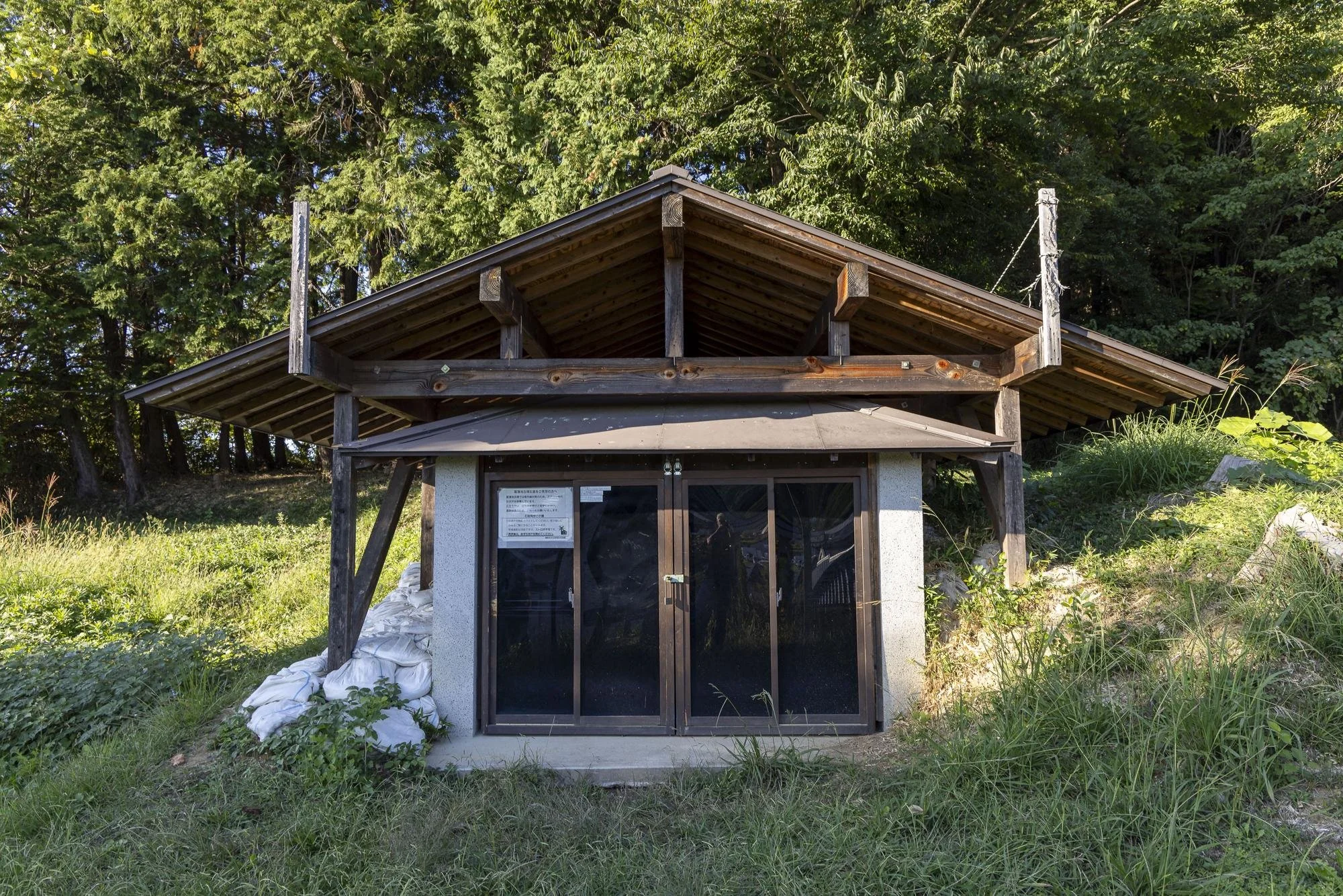 A small rustic building with a wooden shingled roof and glass sliding doors, surrounded by green grass and trees.