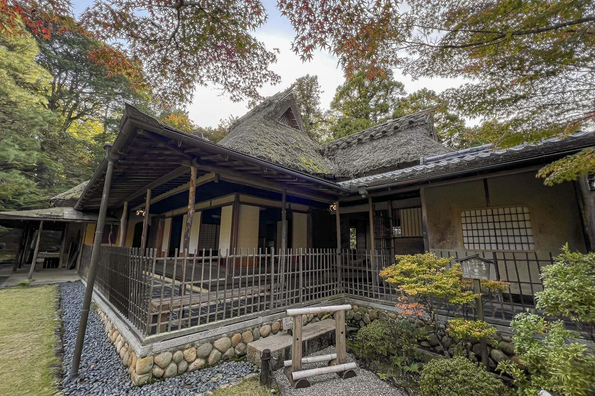 Traditional Japanese building with thatched roof and wooden porch, surrounded by trees and landscaped garden.