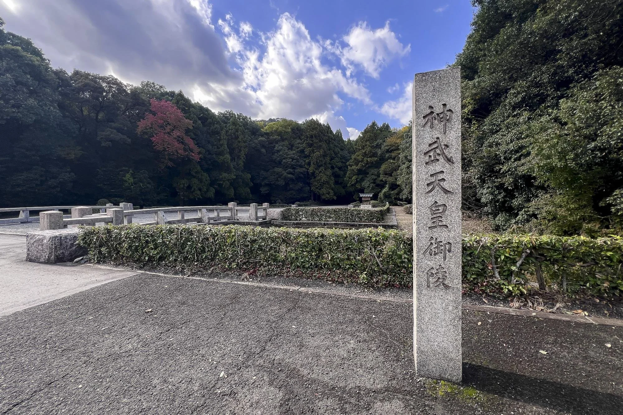 A stone monument with Japanese characters engraved on it, surrounded by lush green trees and a cloudy sky in the background, at a historic site or shrine.