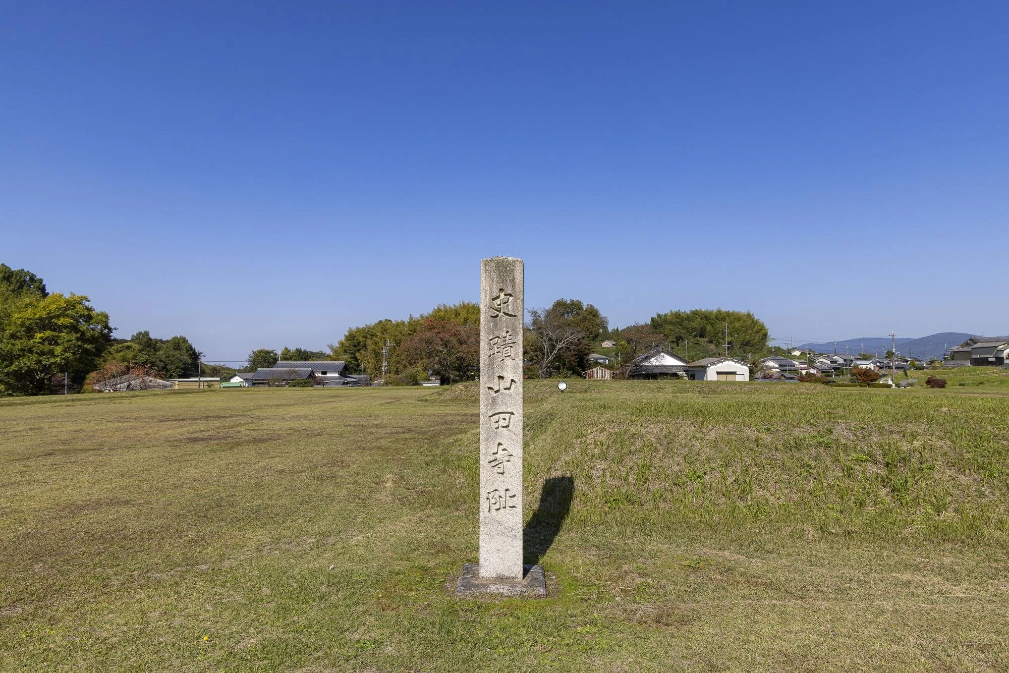 Yamada-dera Temple Site: A stone monument with Japanese inscriptions standing in a grassy field, with a clear blue sky and a small village in the background.