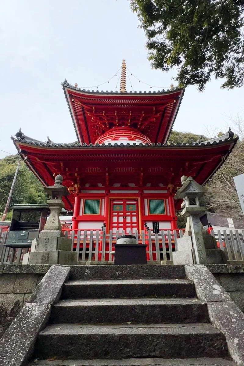 A traditional Japanese shrine with a red wooden structure, stone lanterns on either side, stone steps leading up to it, and a temple bell under the roof. Trees surround the shrine.
