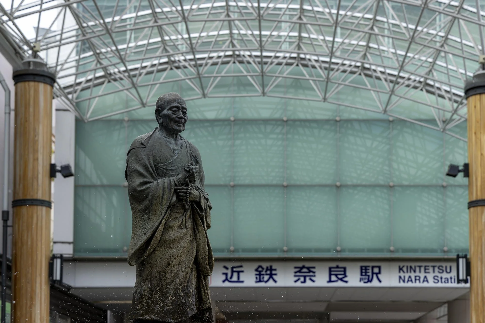 Bronze statue of a smiling elder monk with hands clasped around prayer beads in front of Kintetsu Nara Station in Japan, under a modern glass and steel roof.