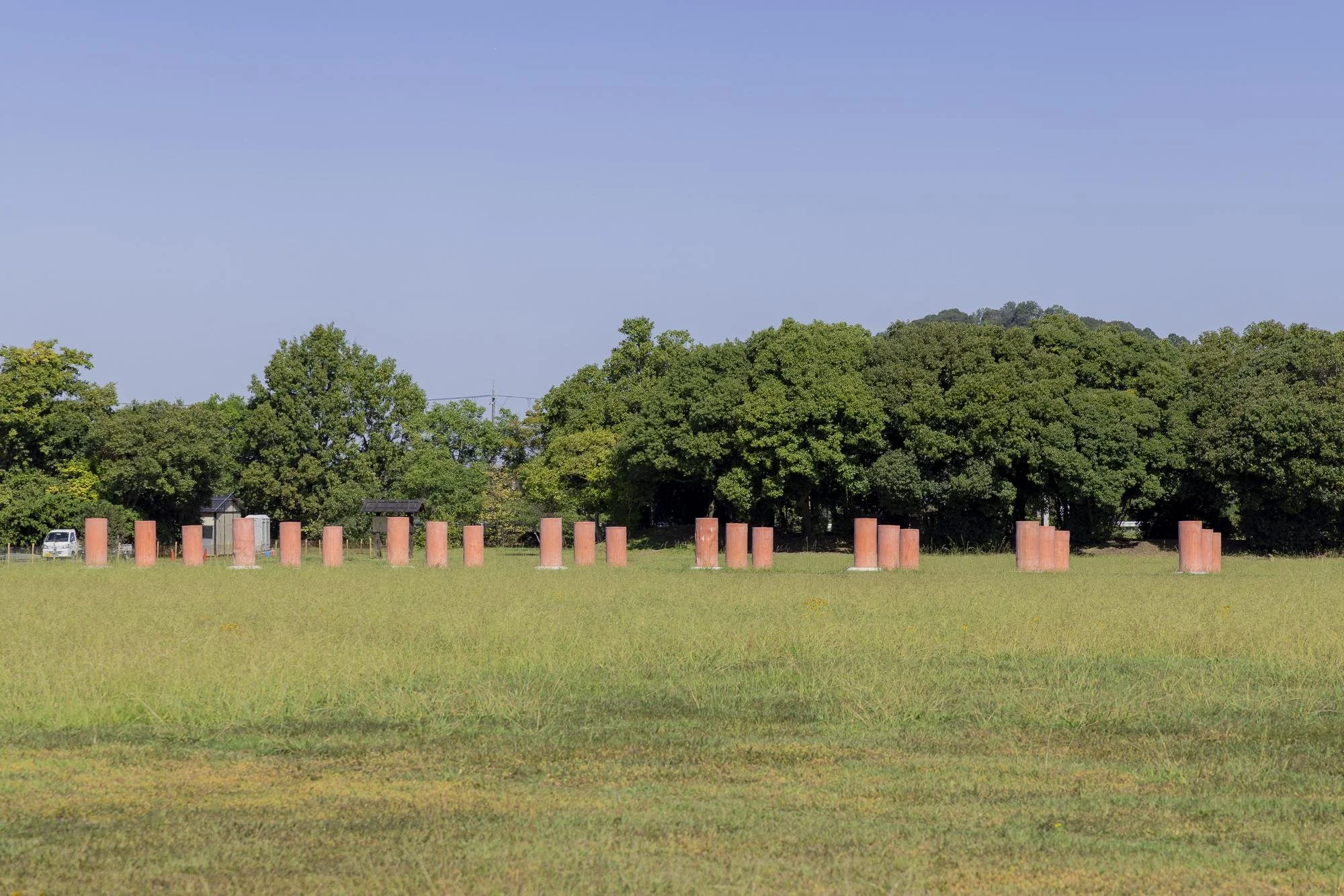Open grassy field with scattered orange cylindrical structures, dense green trees in background, and a clear blue sky.