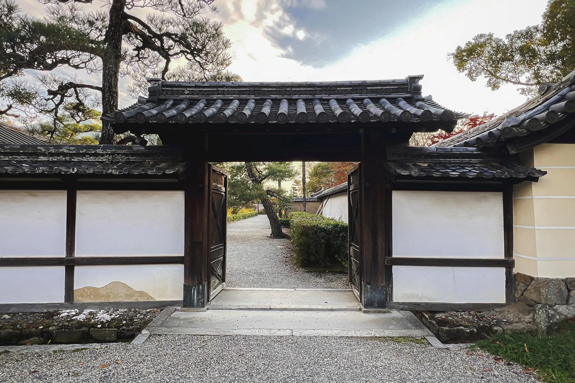 Traditional Japanese wooden gate with tiled roof, leading into a garden with trees and bushes, under a partly cloudy sky.