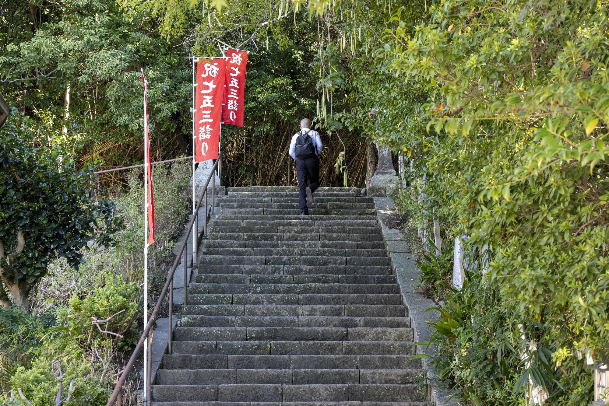 A man with a backpack walking up stone steps surrounded by green foliage and red banners with white Japanese writing.