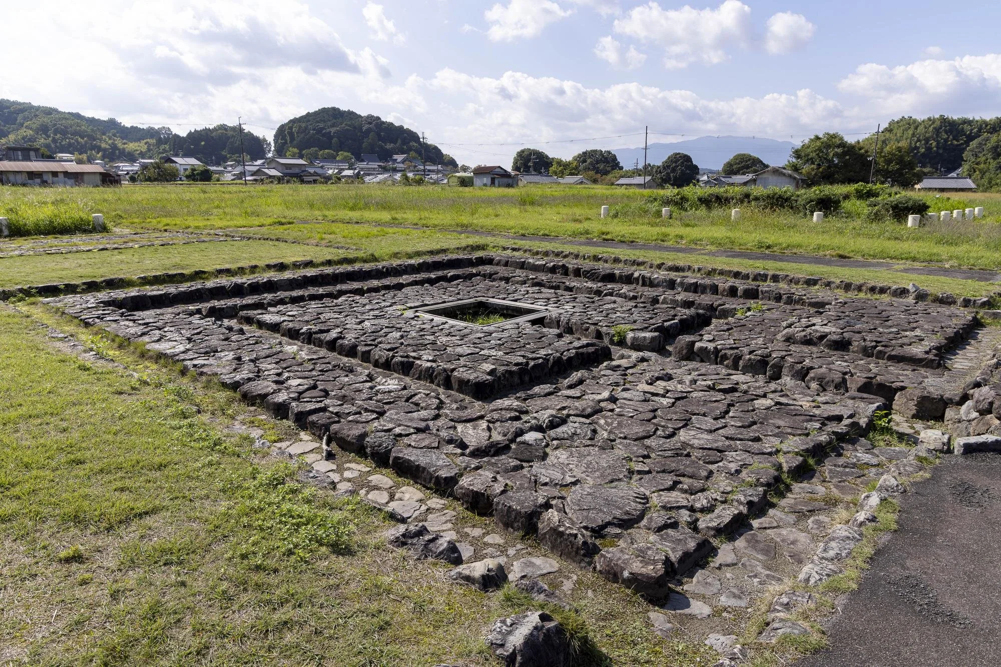 Asuka Palace Site: Ancient stone ruins in a grassy landscape with houses and hills in the background under a partly cloudy sky.
