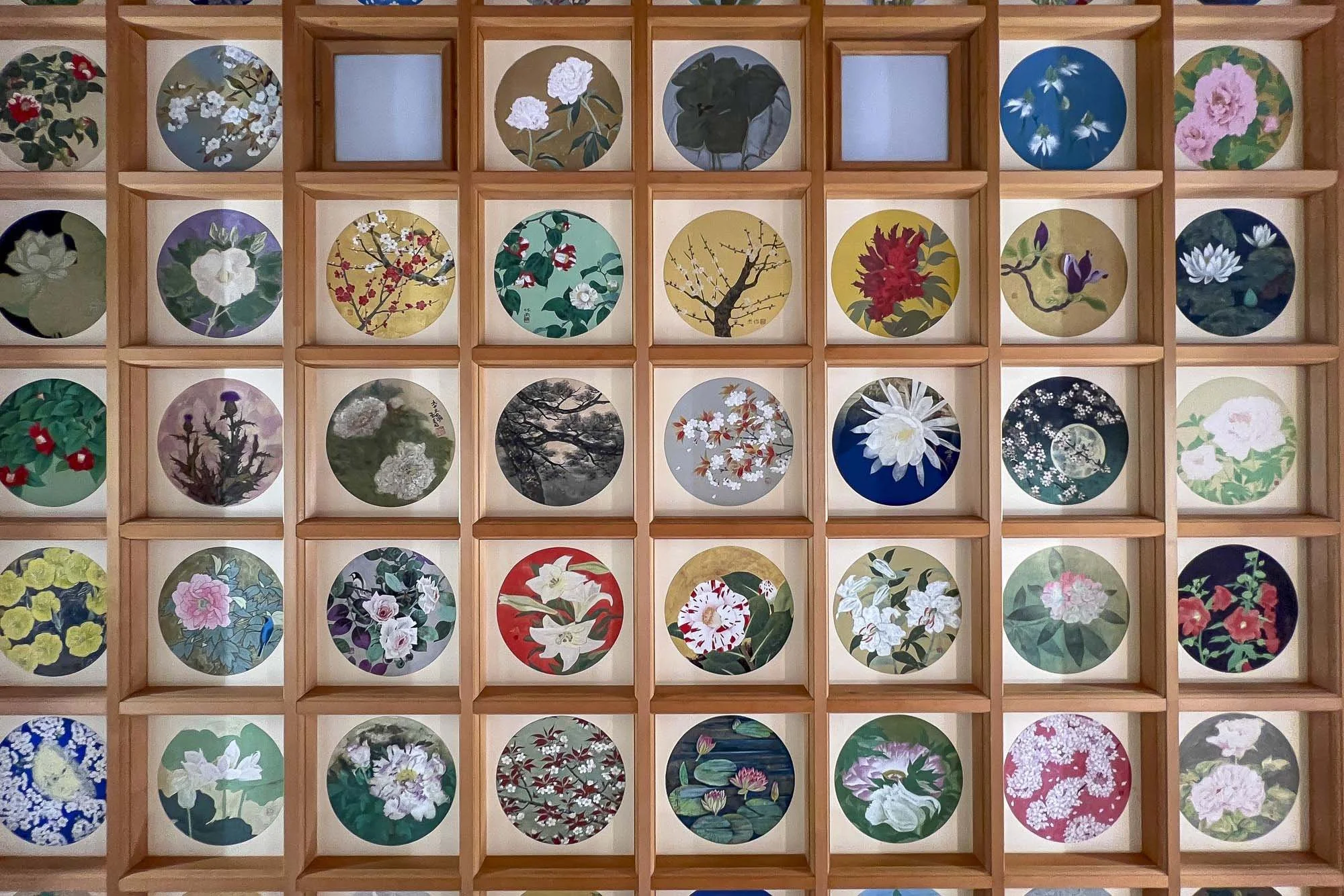 A wooden display shelf with multiple small circular framed paintings of various flowers and nature scenes.