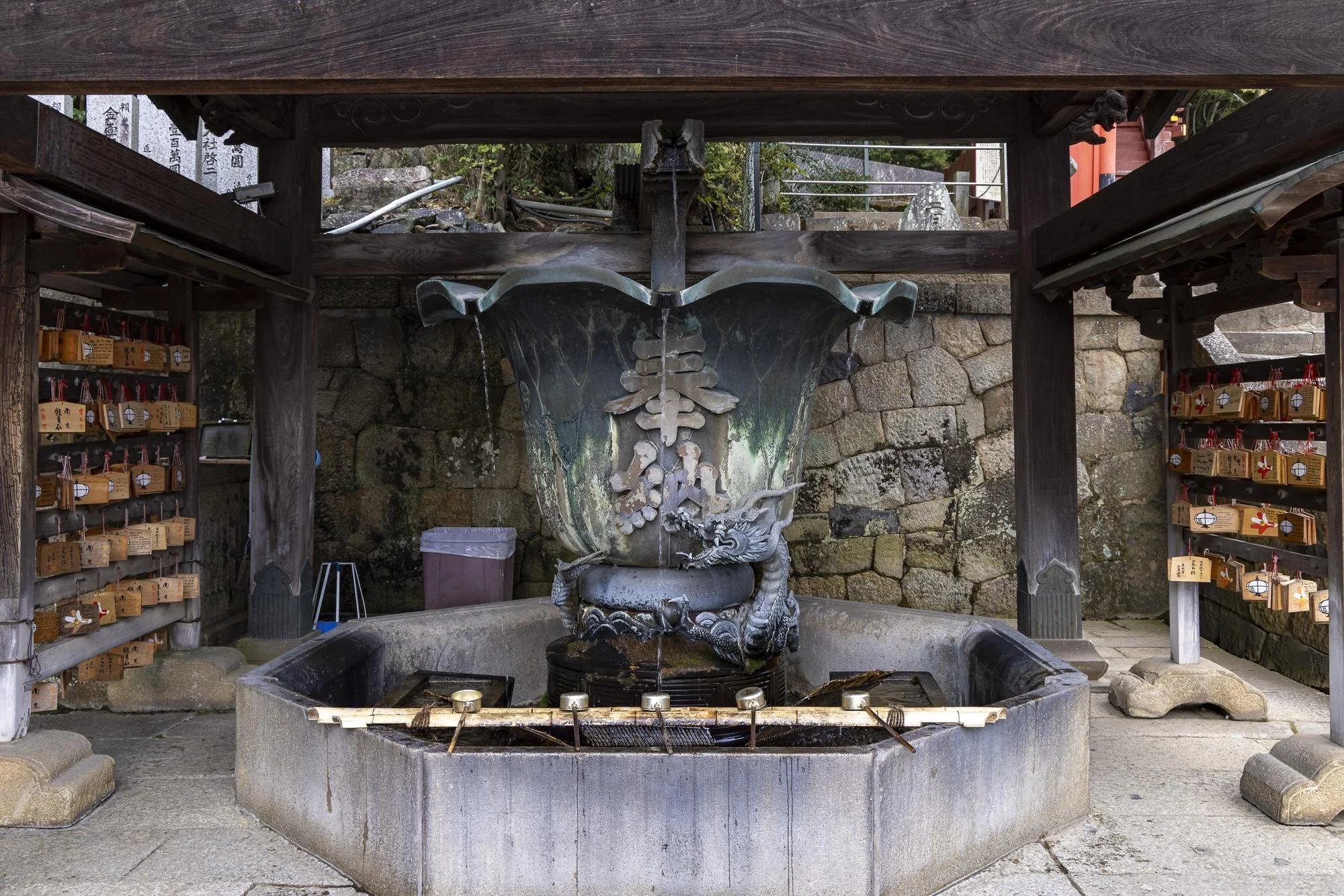 A traditional Japanese water fountain with a dragon sculpture, located under a wooden structure at a shrine, with wooden plaques hanging on both sides.