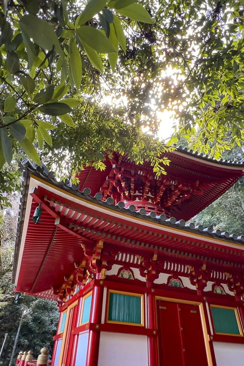 A traditional Japanese pagoda painted in red with colorful accents, situated under green foliage with sunlight filtering through the trees.