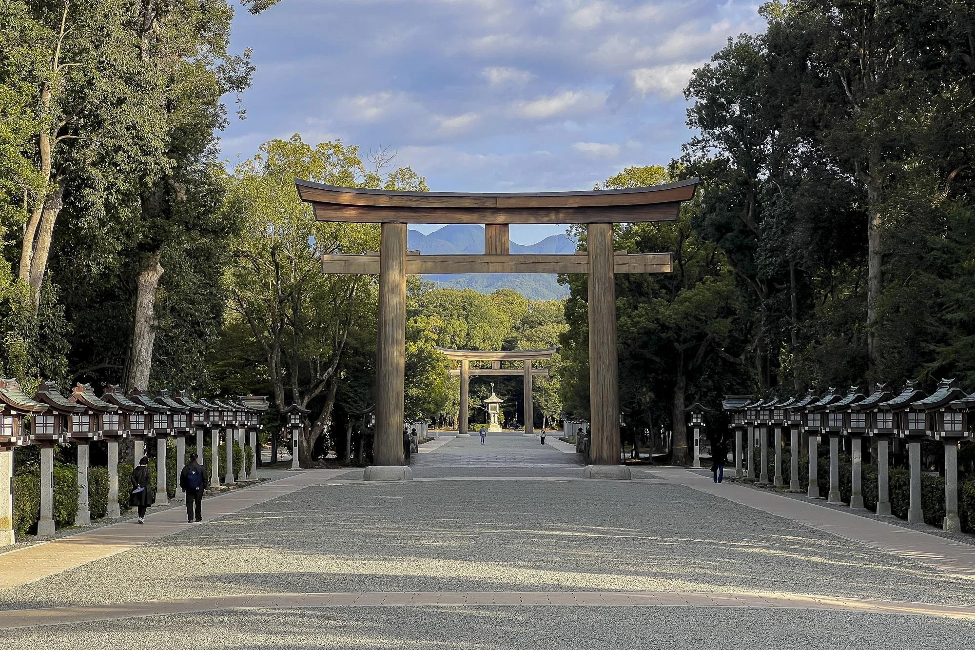 A traditional Japanese torii gate made of wood stands in a pathway surrounded by trees, leading to a shrine with more torii gates further ahead. Mountain peaks are visible in the distance.