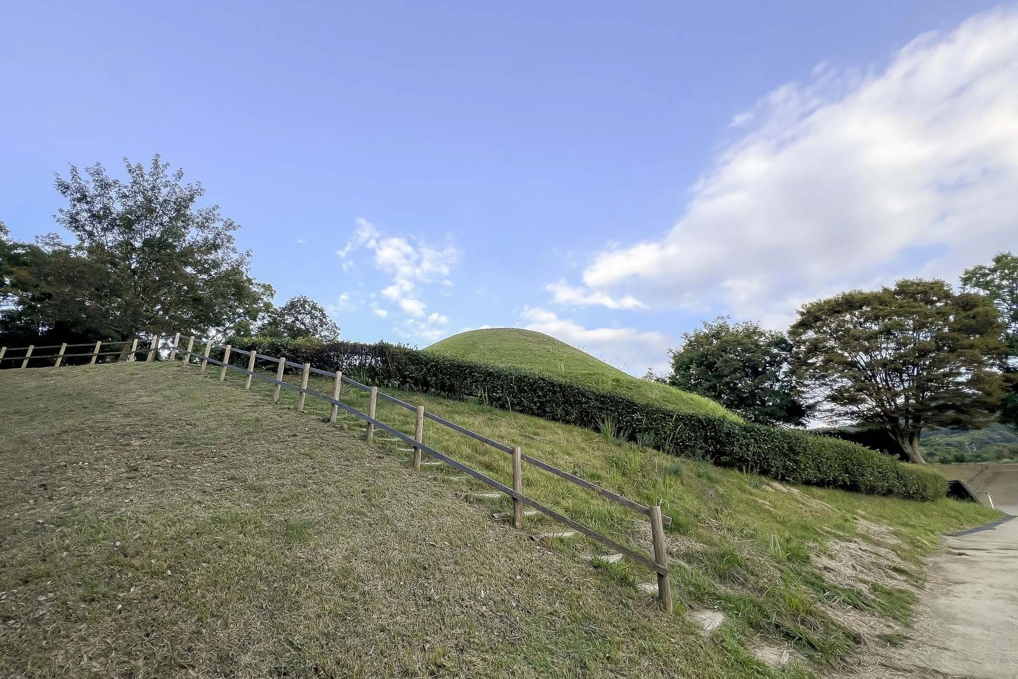 A grassy hillside with a wooden fence and neatly trimmed bushes, with trees on either side and a partly cloudy sky overhead.