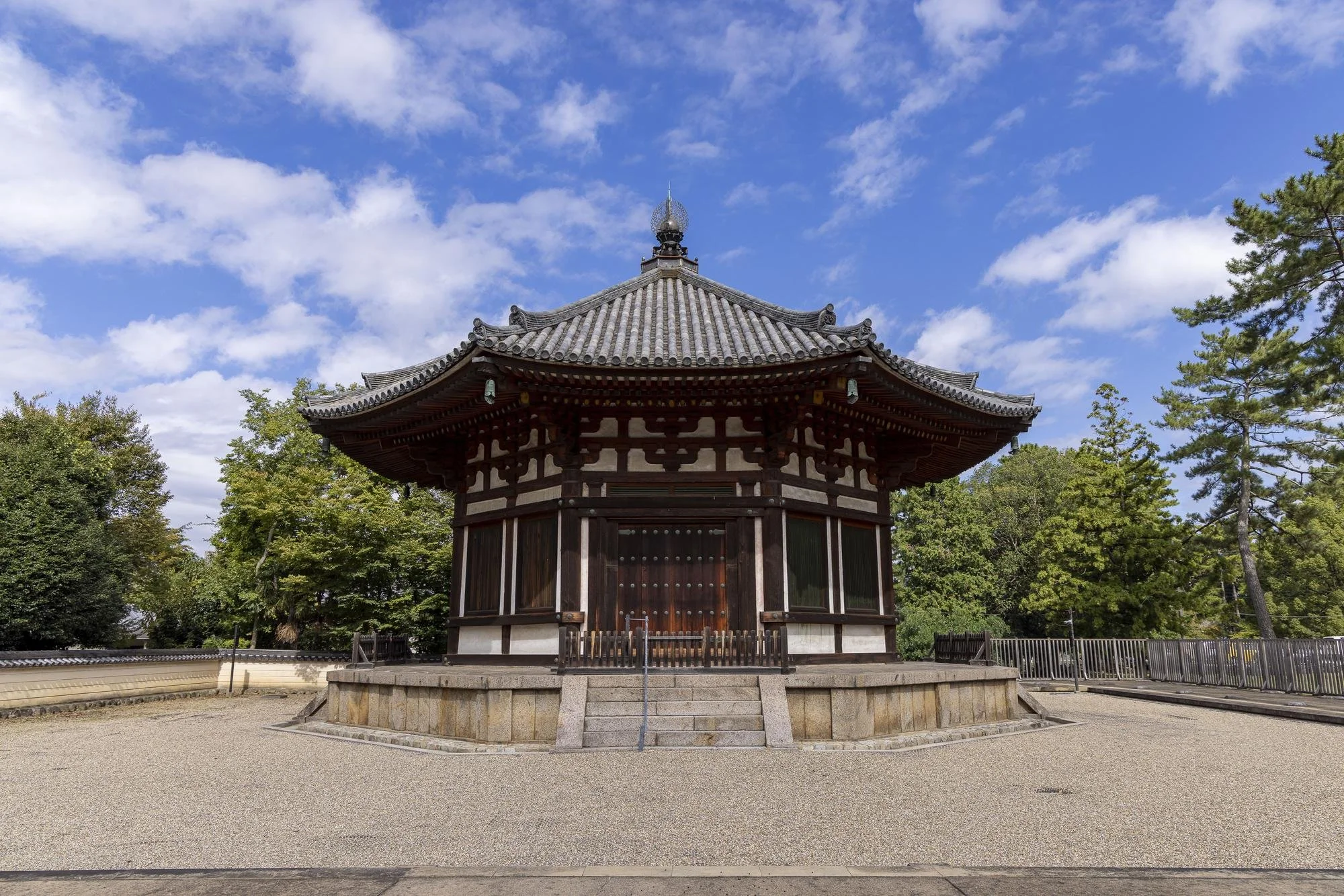 A traditional Japanese wooden shrine with a gray tiled roof, situated on a stone platform, surrounded by green trees under a partly cloudy blue sky.