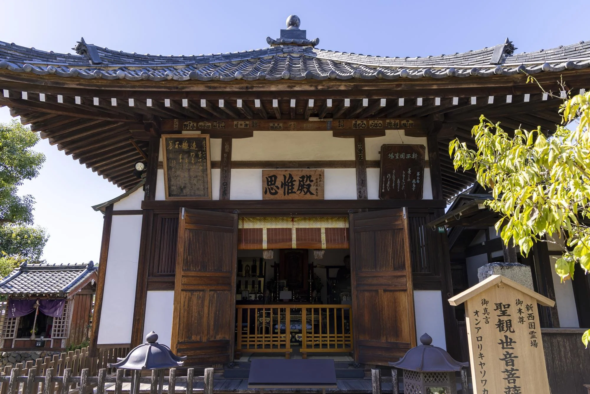 Front view of a traditional Japanese temple with wooden sliding doors, signs with Japanese characters, and a signboard to the right, surrounded by trees under a clear blue sky.