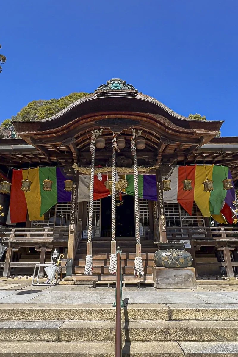 A traditional Japanese shrine with colorful flags hanging in front, ropes with tassels hanging from the roof, and lanterns, set against a blue sky.