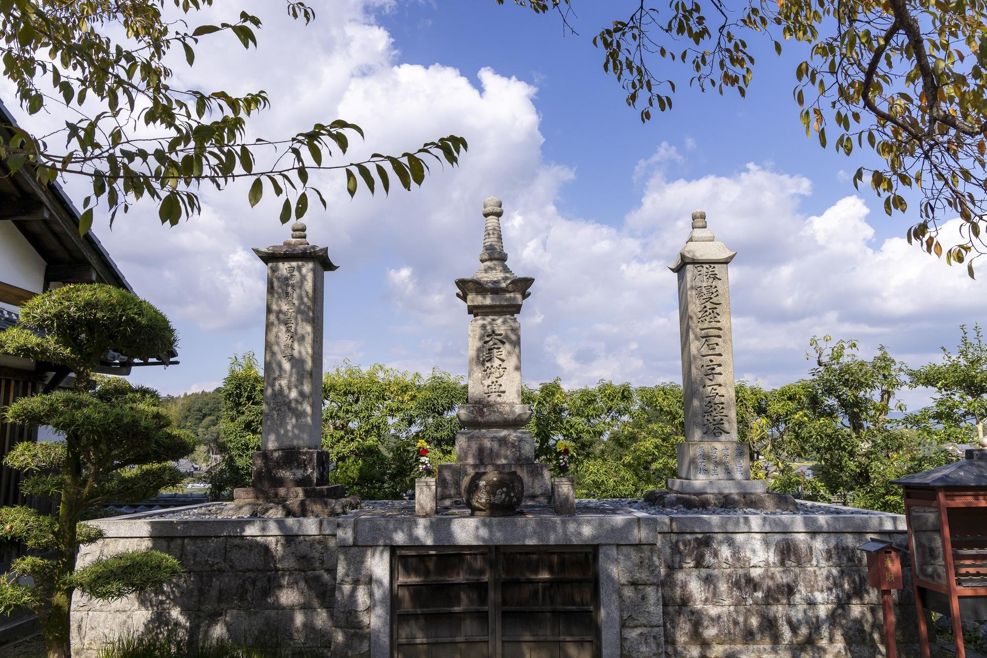 Three stone memorial monuments with tall, narrow, engraved columns are positioned on a stone platform outdoors, with a background of green trees and a partly cloudy sky.