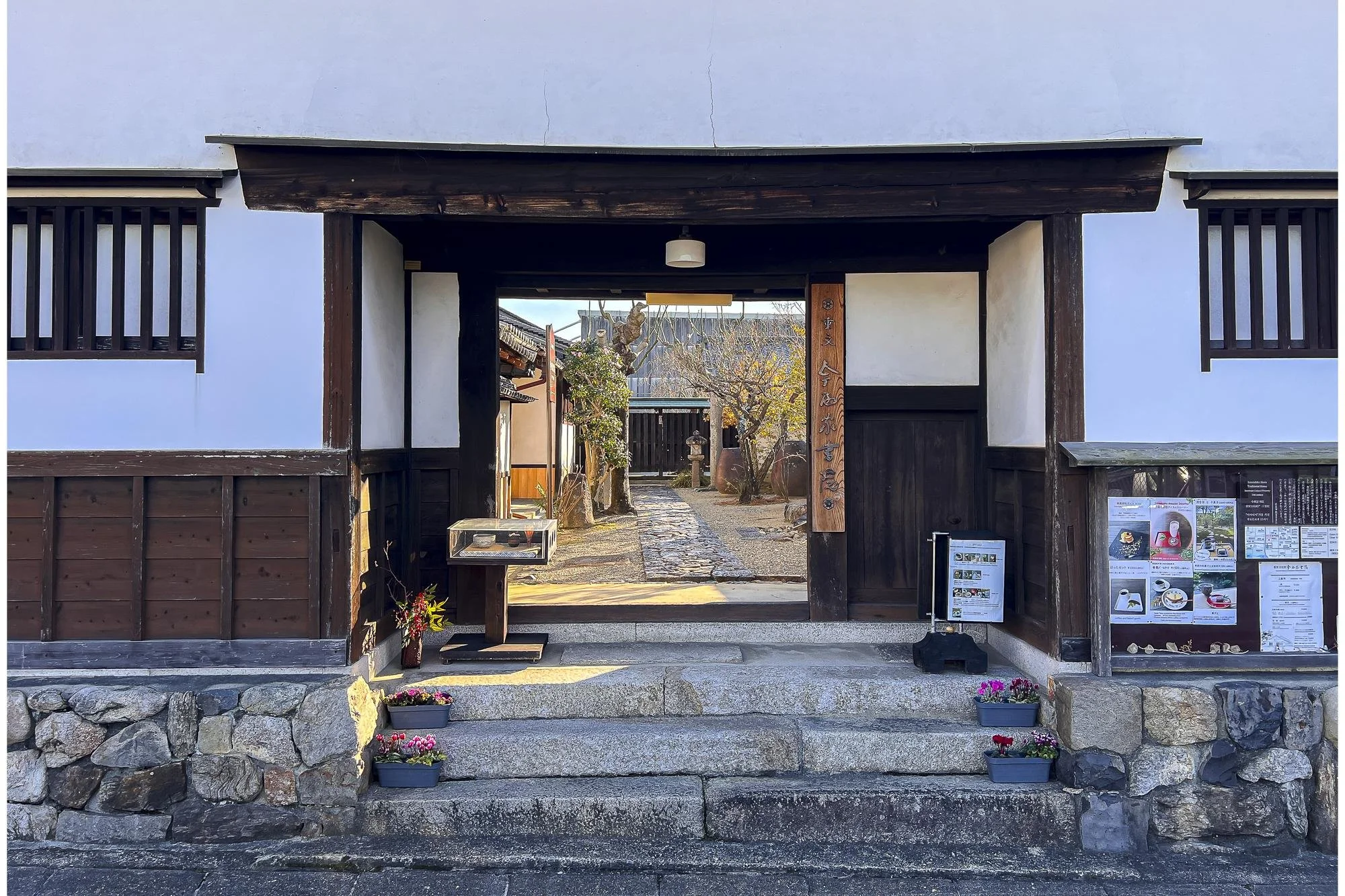 Entrance to a traditional Japanese building with stone steps, wooden accents, and a garden visible through the gate, decorated with potted flowers.