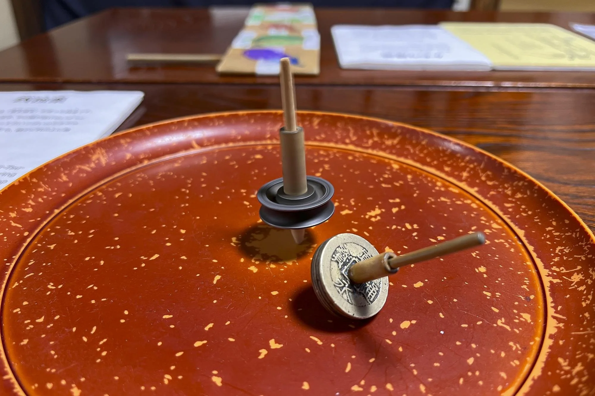 Two Chinese chopsticks standing upright in a red speckled Lazy Susan with two coins on top, on a wooden table with menus and paper in the background.