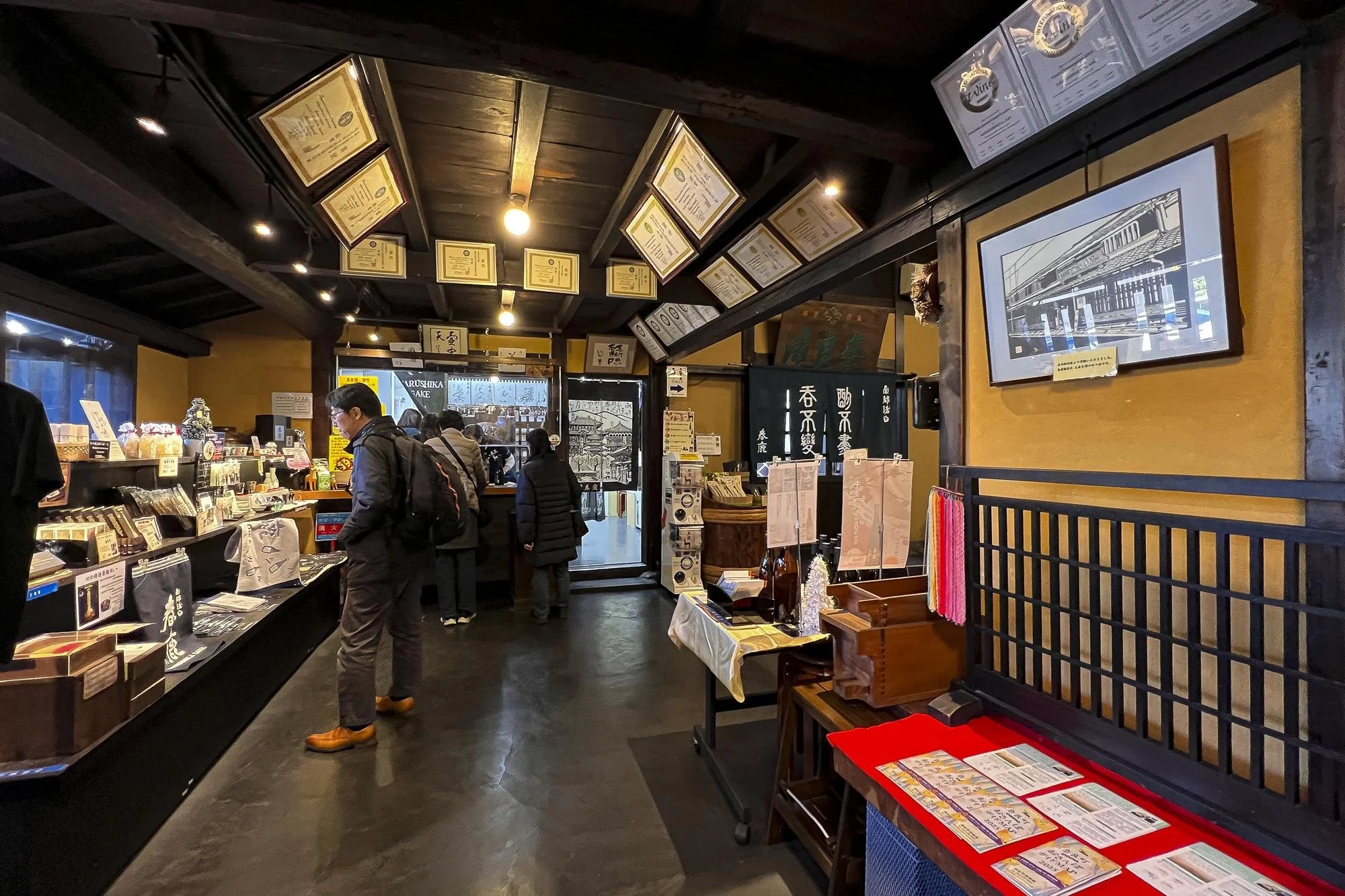 Interior of a Japanese shop or restaurant, with customers browsing and browsing merchandise, dark wood decor, certificates hanging from the ceiling, and displays on counters.