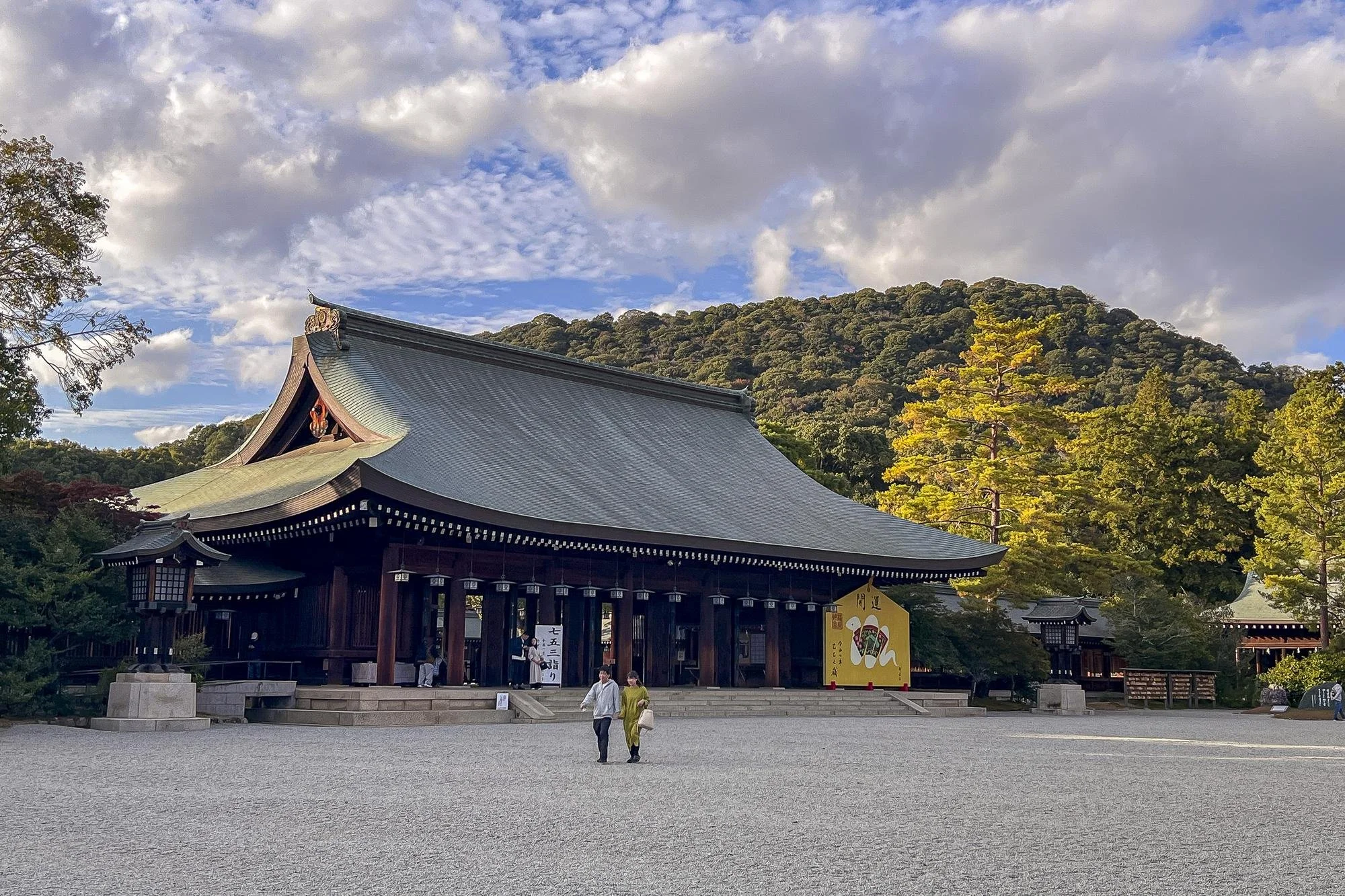 Traditional Japanese shrine with a curved roof, wooden structure, and lanterns, set against lush trees and mountains under a partly cloudy sky.
