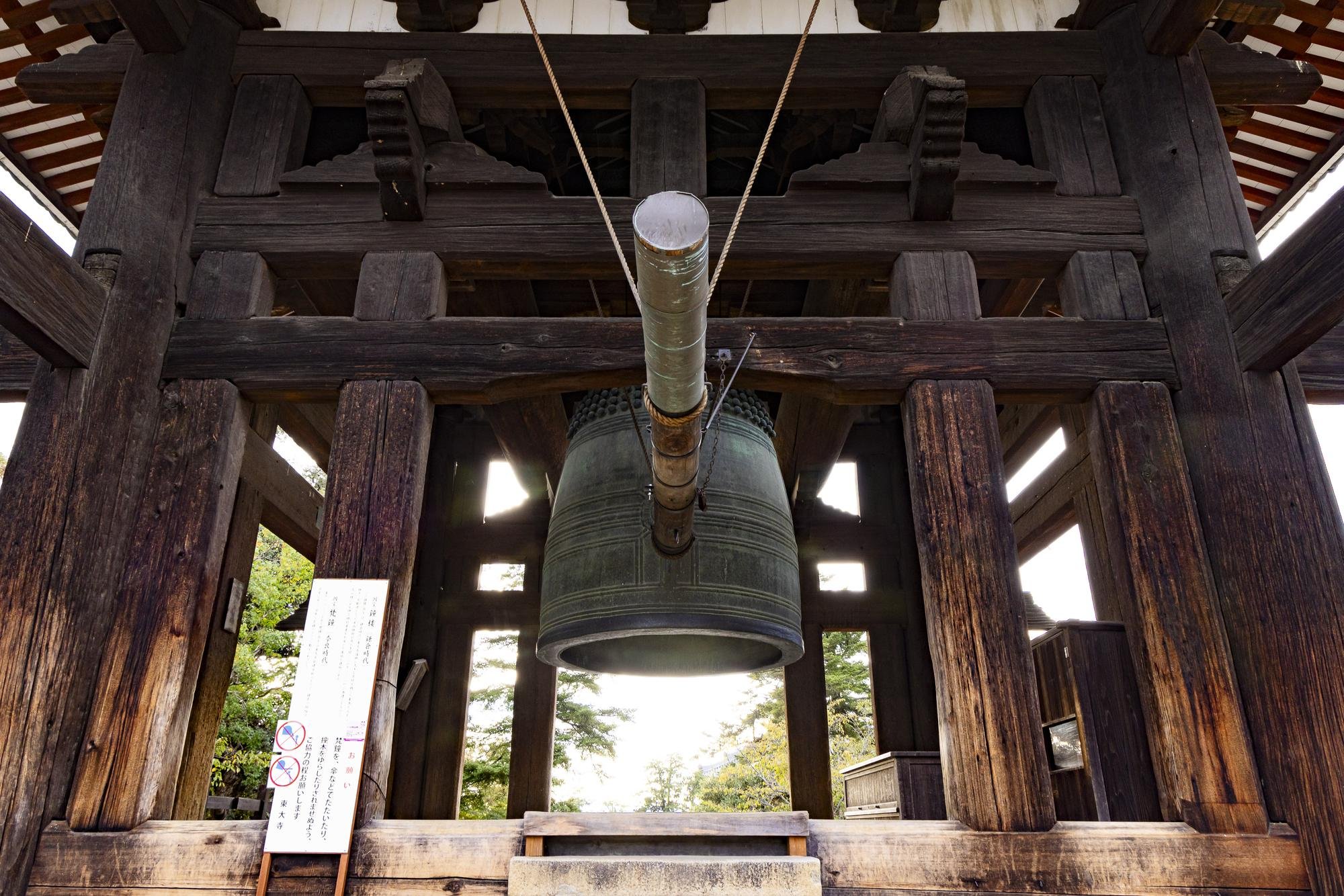 A large bronze bell hanging inside a traditional Japanese wooden structure, with the view from below showing the bell's suspension and the wooden beams supporting it.