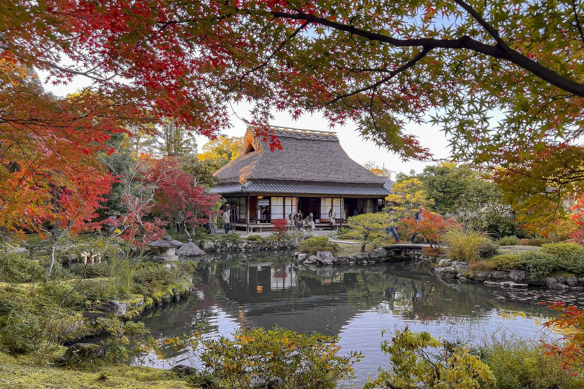 Traditional Japanese house with a thatched roof surrounded by colorful autumn foliage and a pond in the foreground.