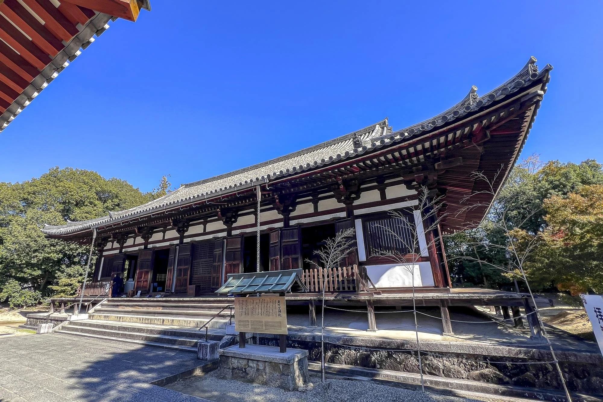 Traditional Japanese temple building with wooden structure, curved tiled roof, and steps leading up to entrance, surrounded by trees with some autumn foliage.