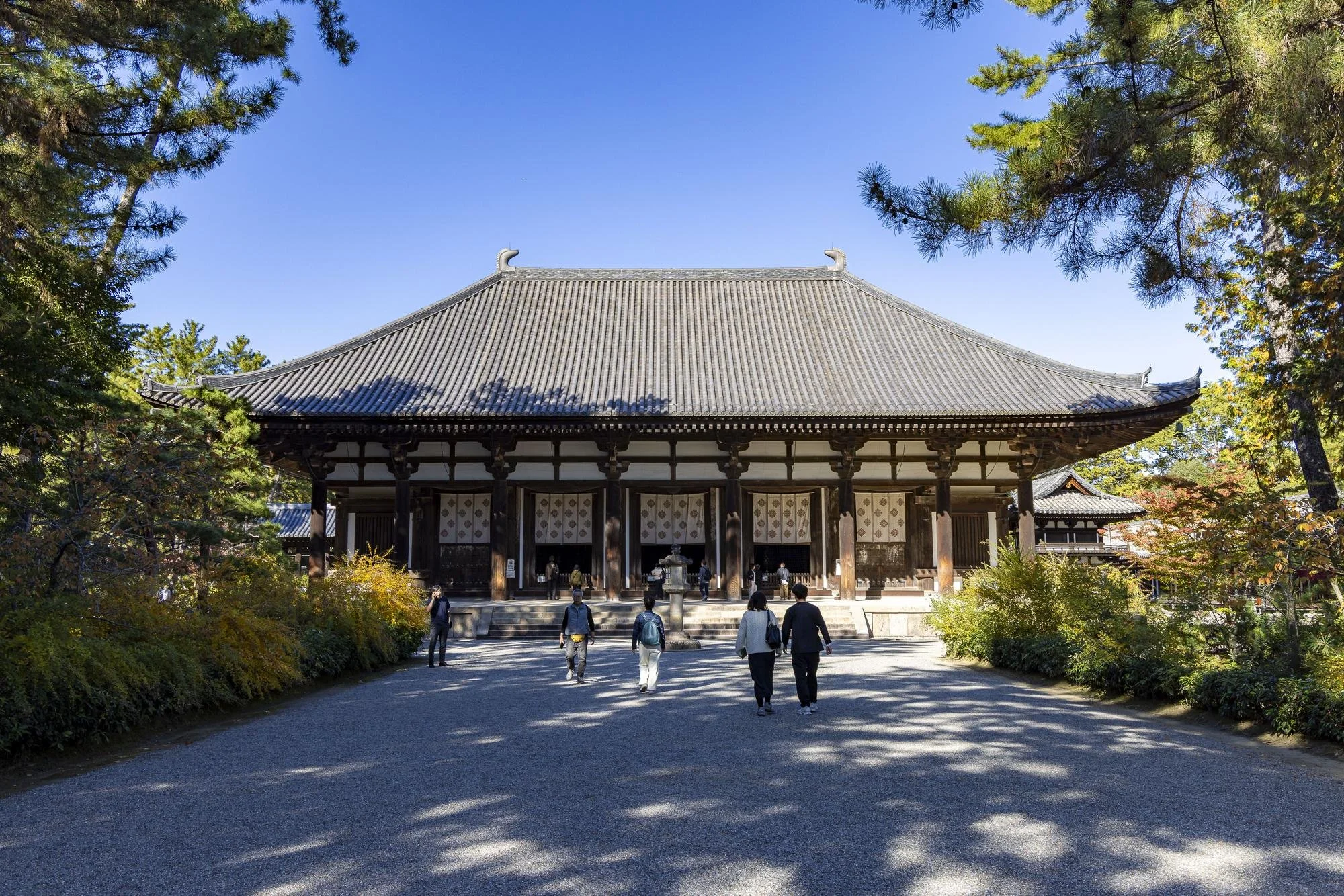 People walking toward a traditional Japanese wooden building with a large, sloped tiled roof, surrounded by trees and foliage on a sunny day.