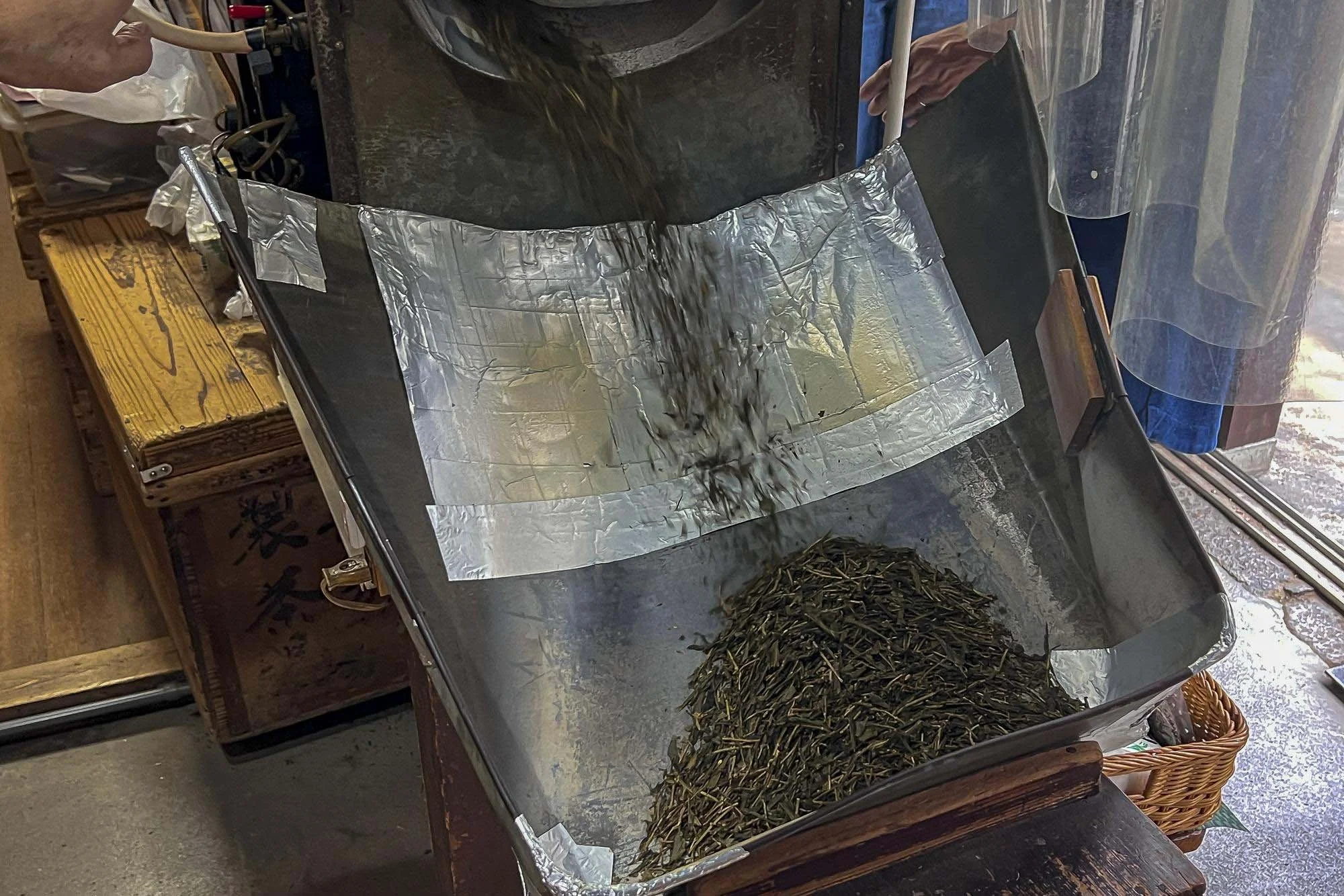 Tea leaves are being poured into a metal container lined with aluminum foil, with some leaves already inside, in a tea shop or processing area.