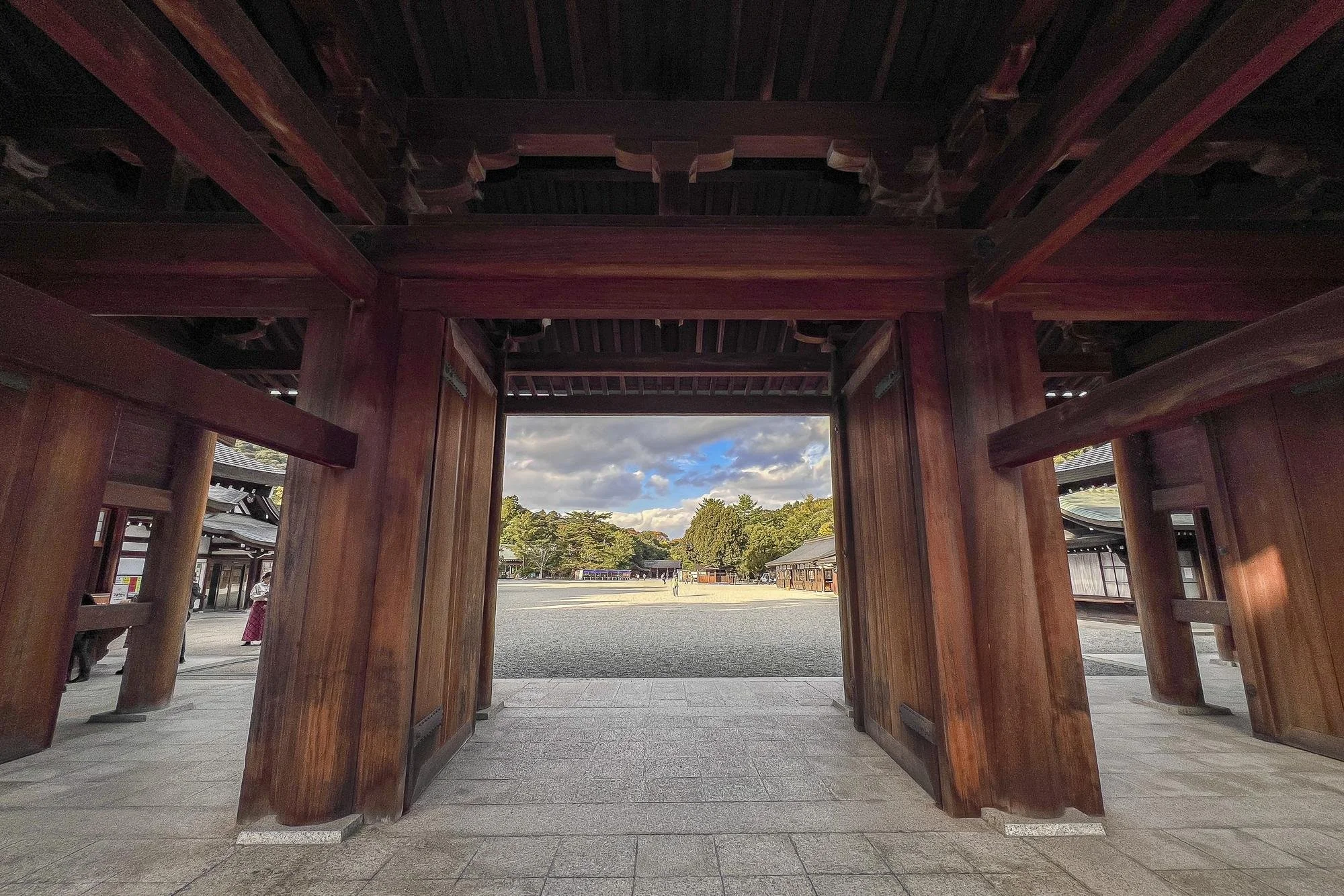 View through a wooden gate at a traditional Japanese shrine with trees, buildings, and a cloudy sky in the background.