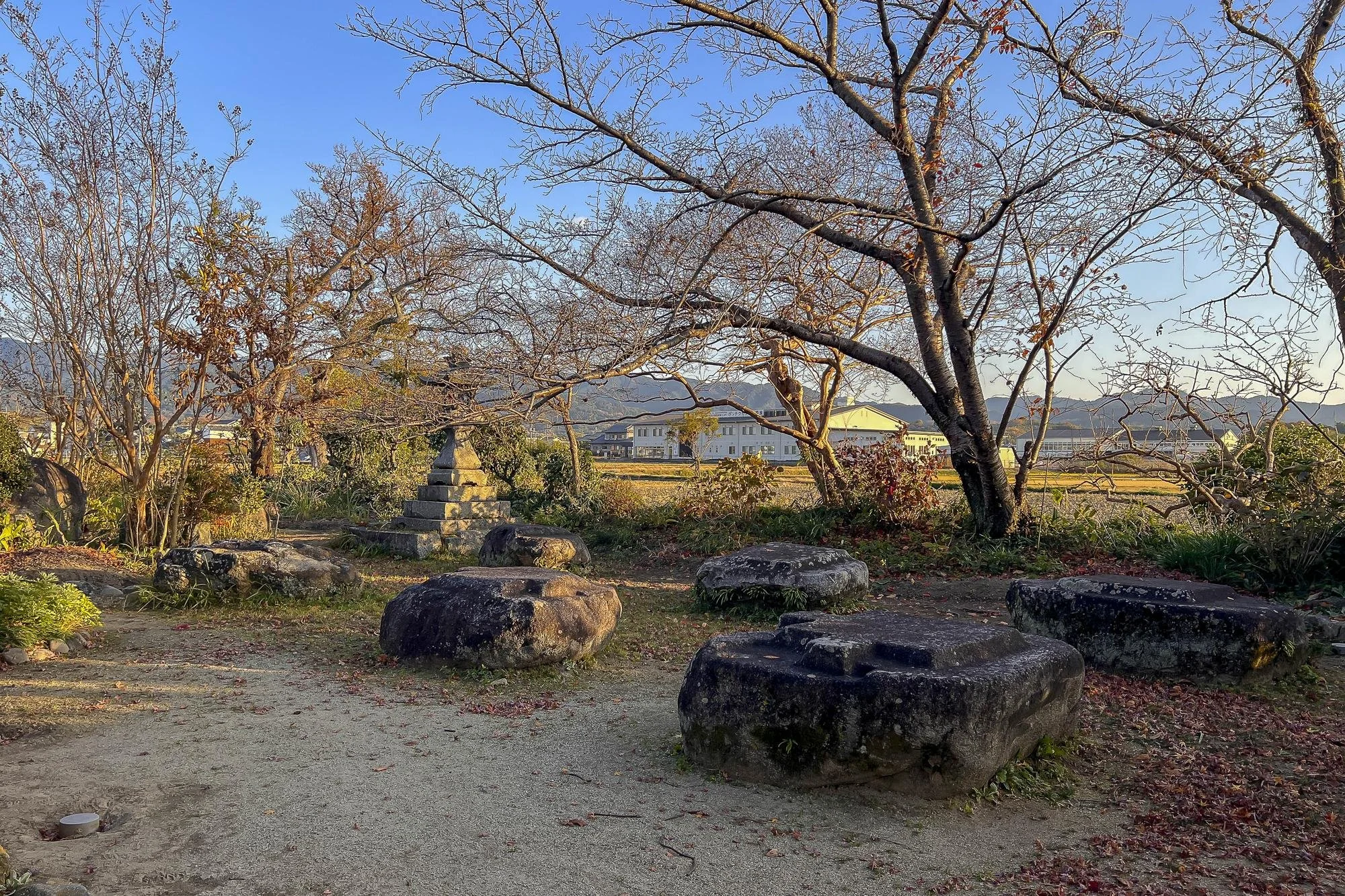 A garden with leafless trees, large rocks, and a stone pagoda monument, with a background of buildings and distant mountains under a clear blue sky.